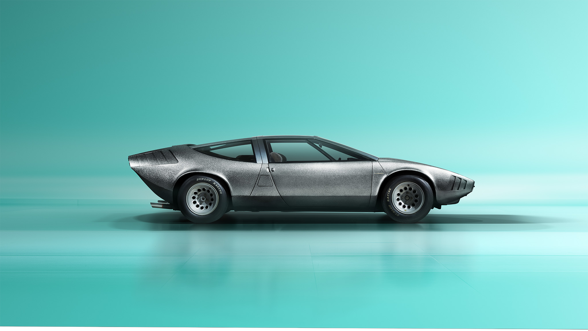 A sleek, vintage Alfa Romeo 33 sports car with a silver body and an aerodynamic design is displayed in profile against a minimalist turquoise background. The car features a low, sharp front end and a uniquely shaped rear. This retouched masterpiece looks like it just rolled off the pages of Magneto Magazine.