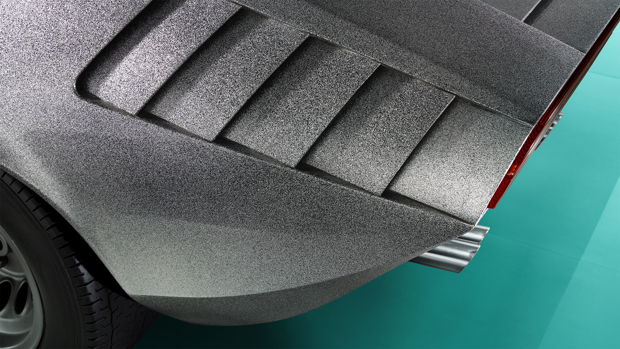 Close-up of the rear portion of a classic silver Alfa Romeo 33 sports car, showing distinctive air vents, a tire, and part of the taillight against a teal background. The angle emphasizes the car's vintage design and unique textured paint—a perfect capture for Magneto Magazine by an expert retoucher.