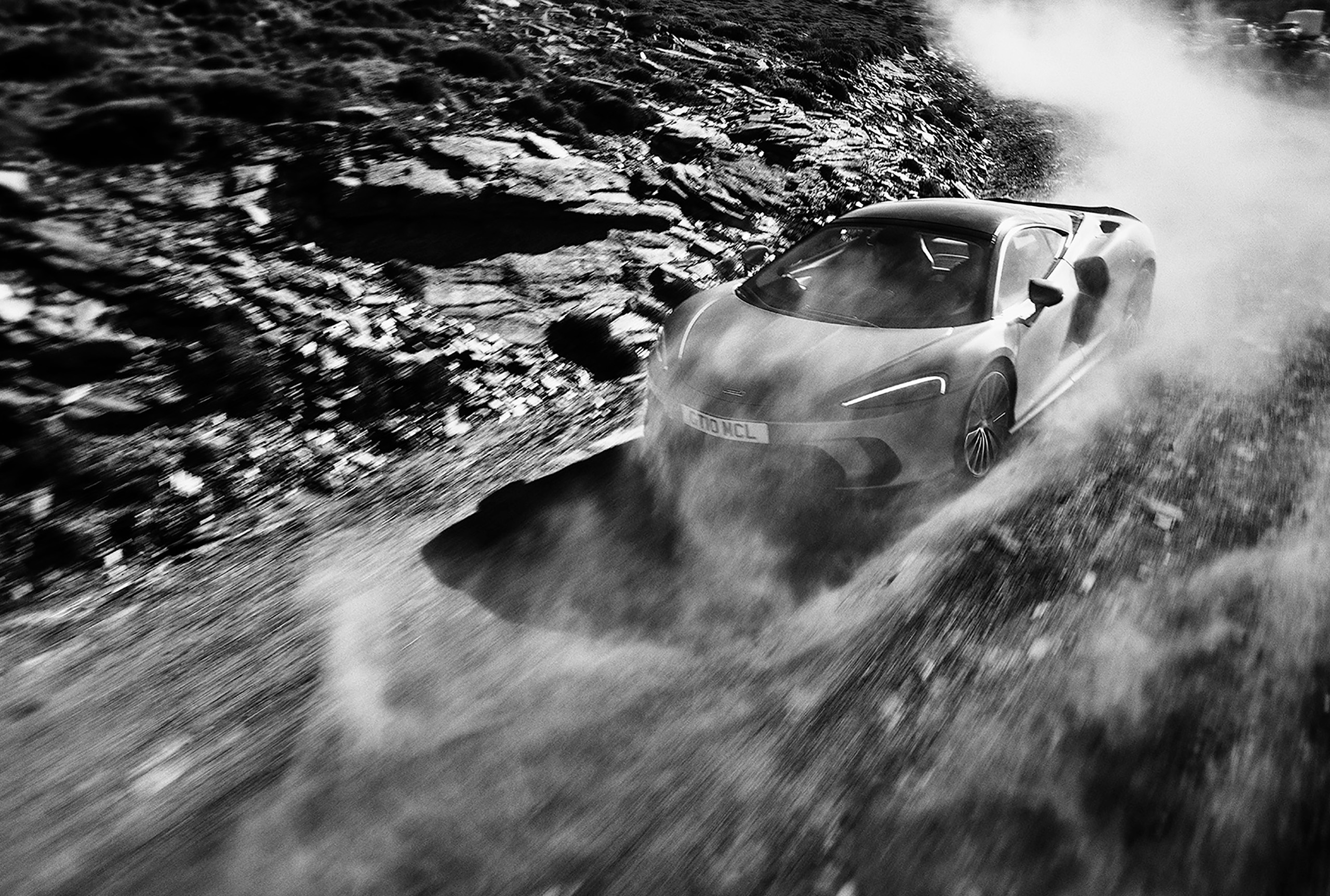 A sleek, modern McLaren grand tourer speeds through a rocky, dusty terrain, leaving a dramatic plume of dust behind it. The black-and-white image captures the motion and intensity of the scene perfectly, with an expert automotive retoucher enhancing the car cutting through the rugged landscape.