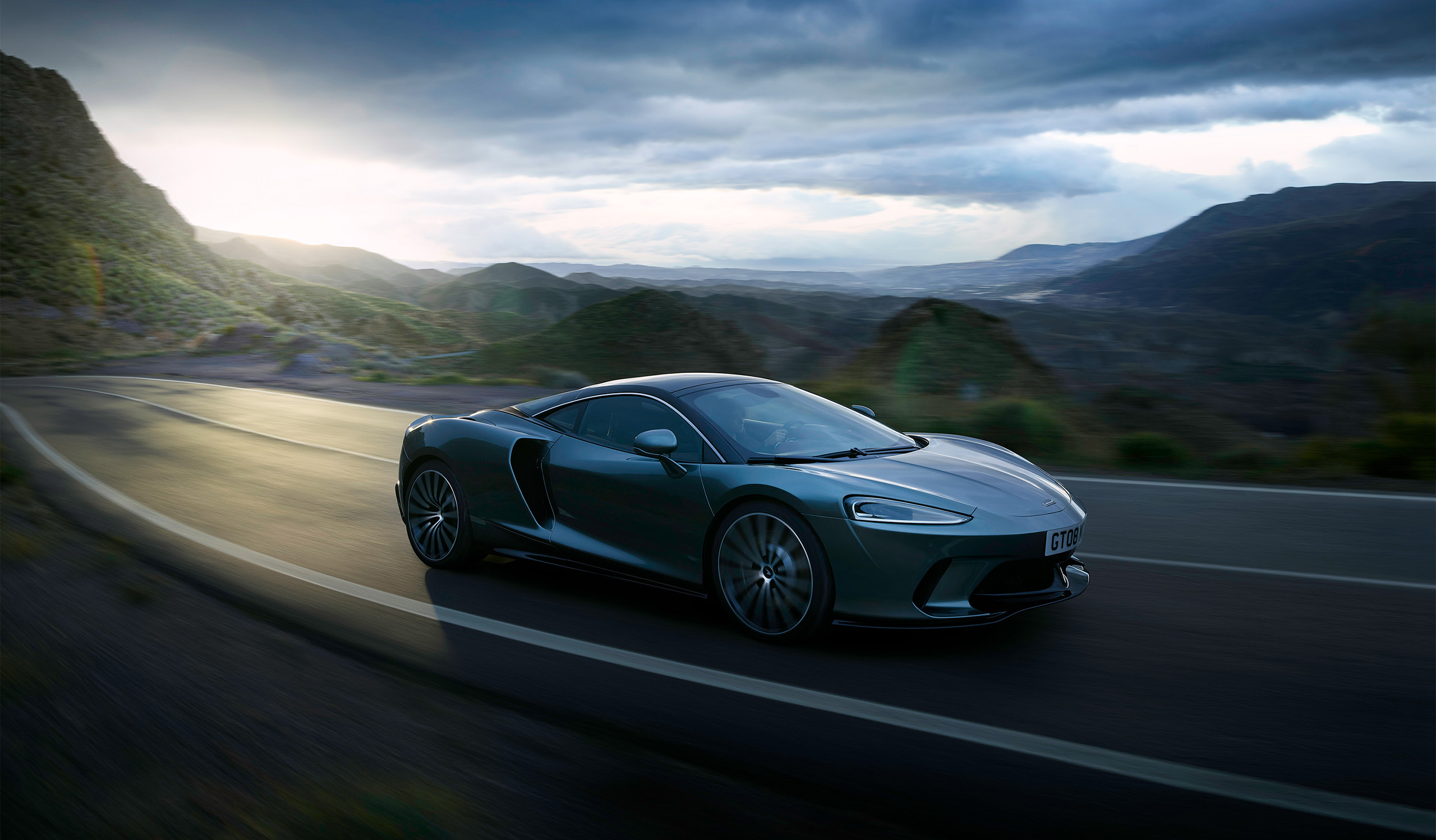 A sleek silver McLaren grand tourer drives on a winding mountain road at dusk. The background features rolling hills and a dramatic, cloudy sky with rays of sun peeking through, creating a dynamic and scenic atmosphere perfect for an automotive retoucher's touch.