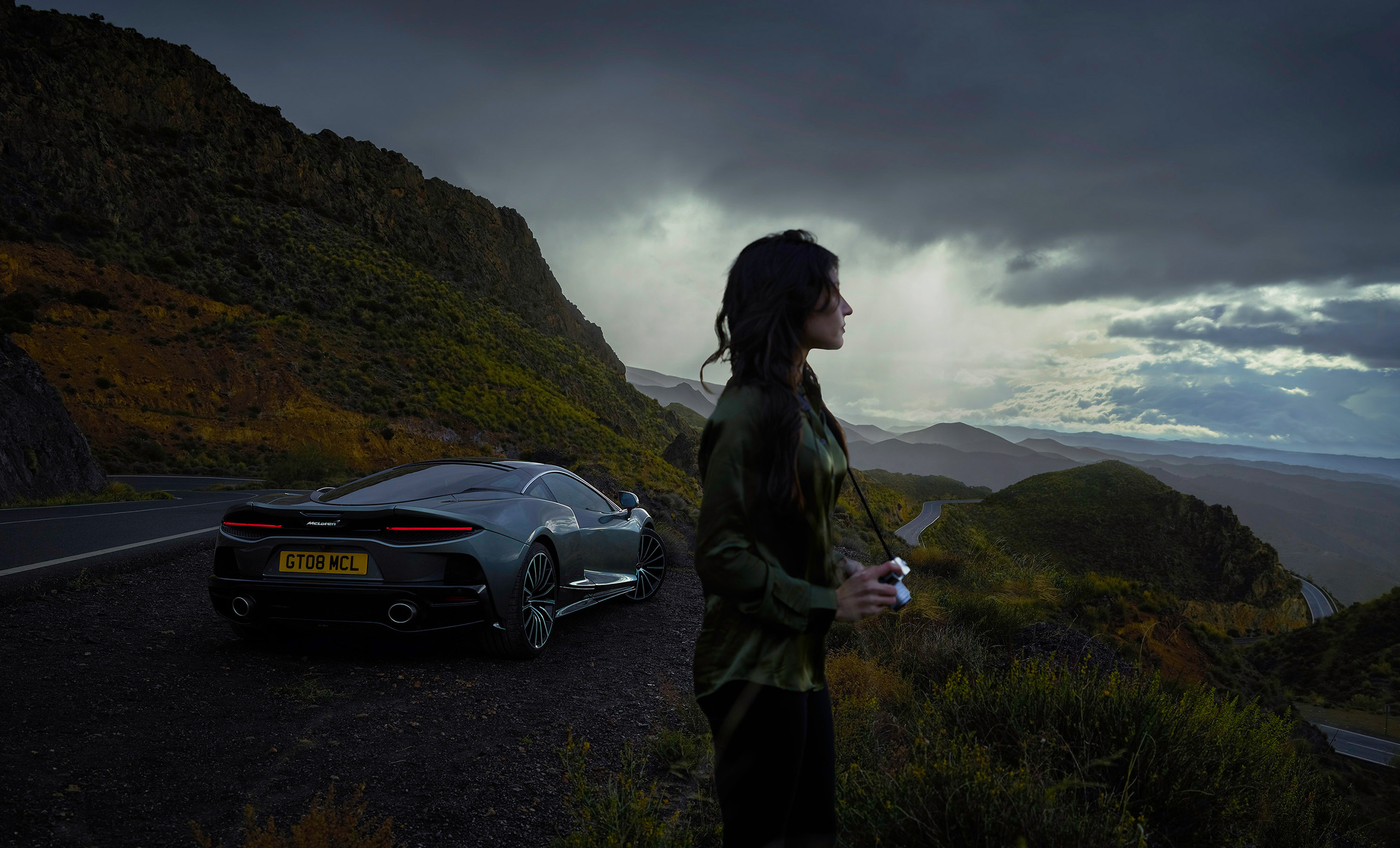 A woman stands holding a camera, gazing at a mountainous landscape under a cloudy sky. Behind her is a parked McLaren Grand Tourer with license plate GT08 MCL on a winding road, perfectly captured by an automotive retoucher. The scene exudes adventure and the raw beauty of nature.