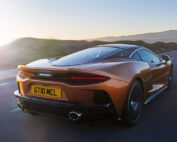 An orange McLaren Grand Tourer GTdrives on a scenic road at sunset, showcasing its sleek design and aerodynamic features. The rear license plate reads "GT10 MCL." The backdrop includes a serene mountainous landscape bathed in soft, golden light. Perfect for any automotive retoucher's portfolio.