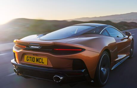 An orange McLaren Grand Tourer GTdrives on a scenic road at sunset, showcasing its sleek design and aerodynamic features. The rear license plate reads "GT10 MCL." The backdrop includes a serene mountainous landscape bathed in soft, golden light. Perfect for any automotive retoucher's portfolio.
