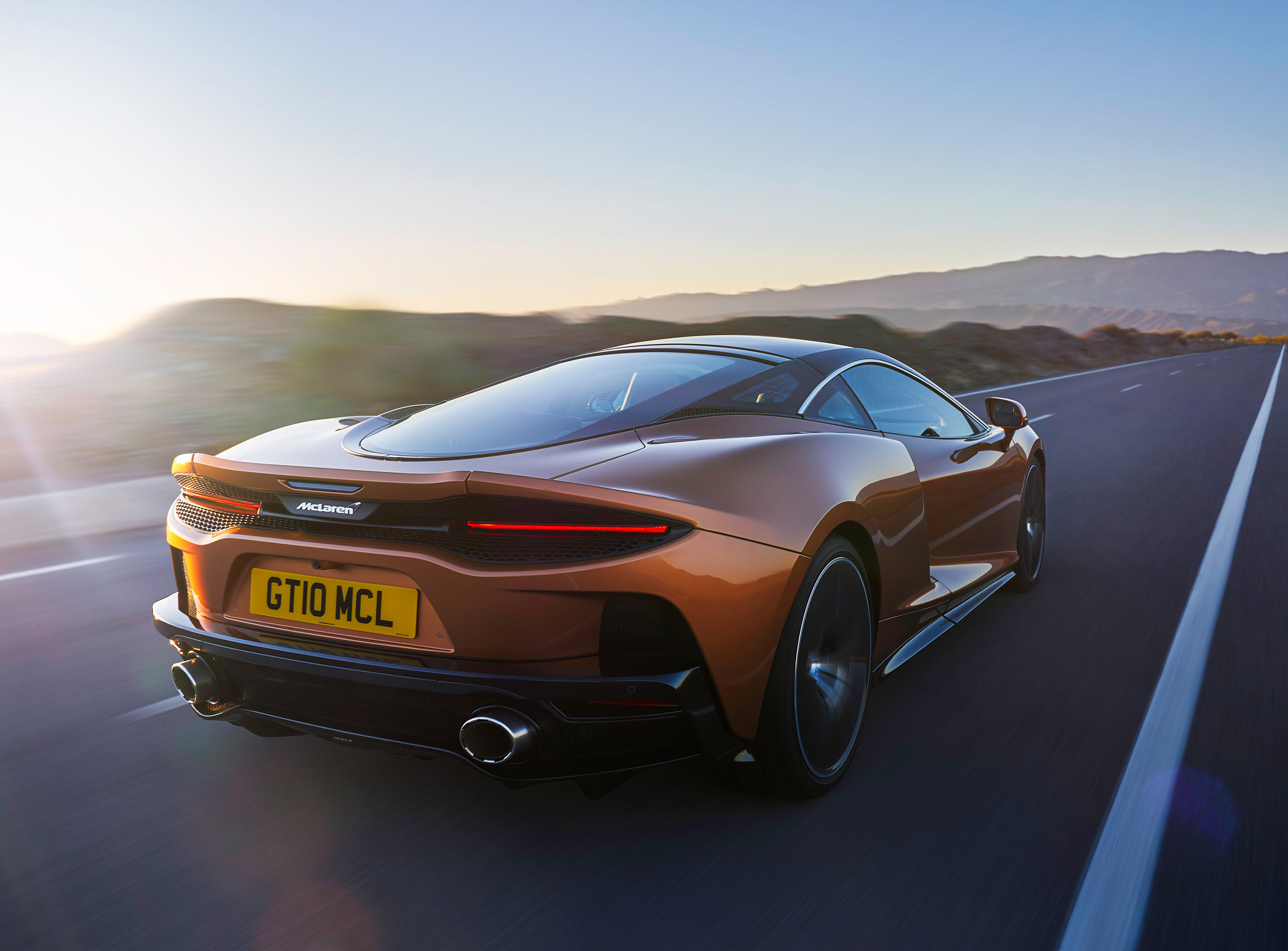 A sleek, orange McLaren grand tourer drives on a scenic highway during sunset. The car, a McLaren GT, has the license plate "GT10 MCL" and features a streamlined design with glowing taillights. An automotive retoucher would appreciate the mountains and clear skies in the background.