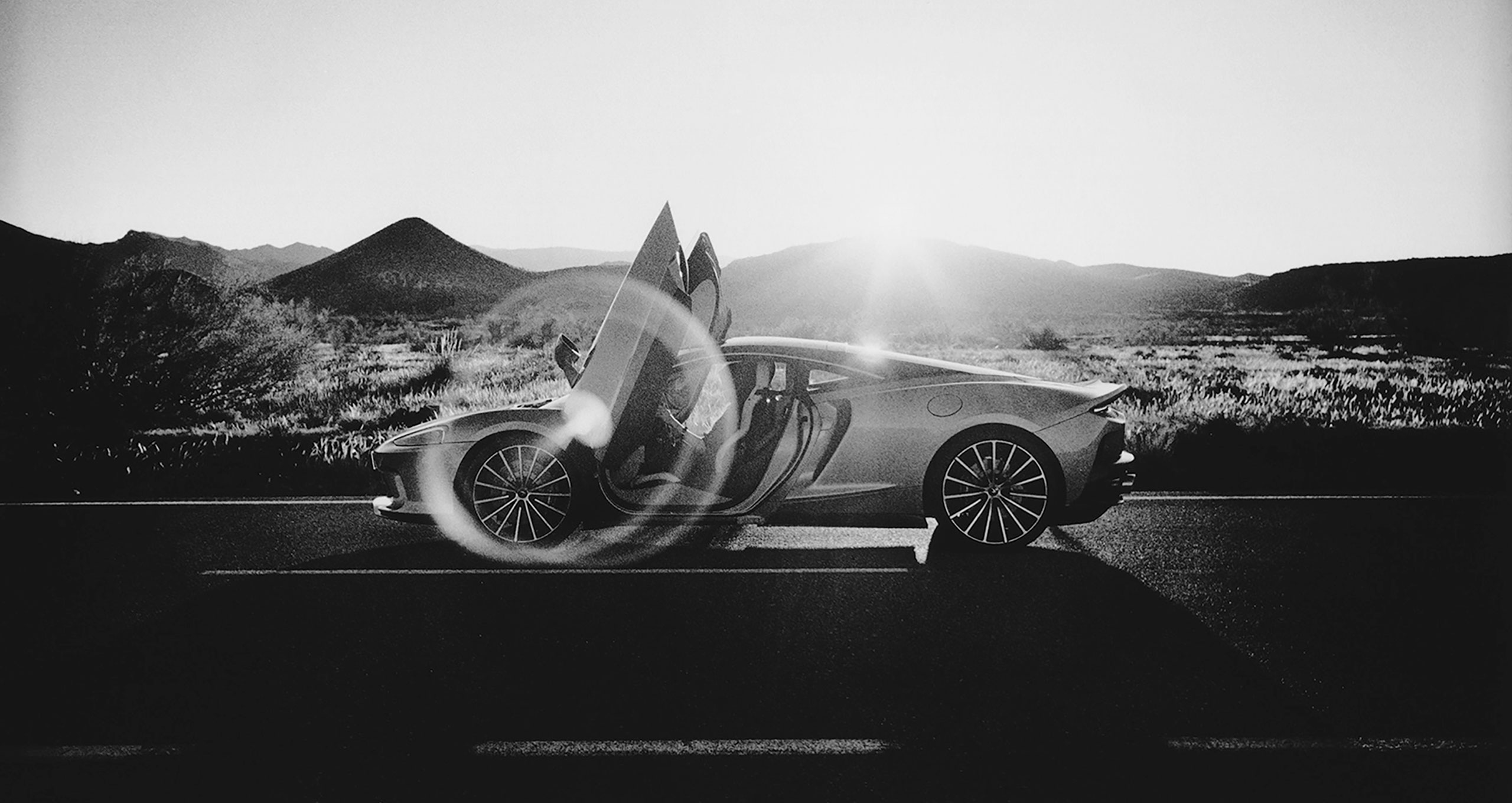 Black and white image of a sleek, modern McLaren grand tourer with butterfly doors open, parked on a road in a mountainous desert landscape. The sun is setting behind the hills, casting a dramatic light over the scene, perfect for an automotive retoucher's dream.