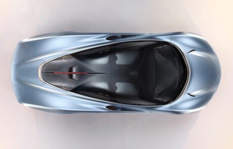 Top view of a sleek, silver-blue McLaren Speedtail with a futuristic design. The car retoucher highlights its streamlined body, transparent roof sections, and smooth curves, showcasing its aerodynamic shape. The interior is minimally visible, with seating and control details faintly discernible.