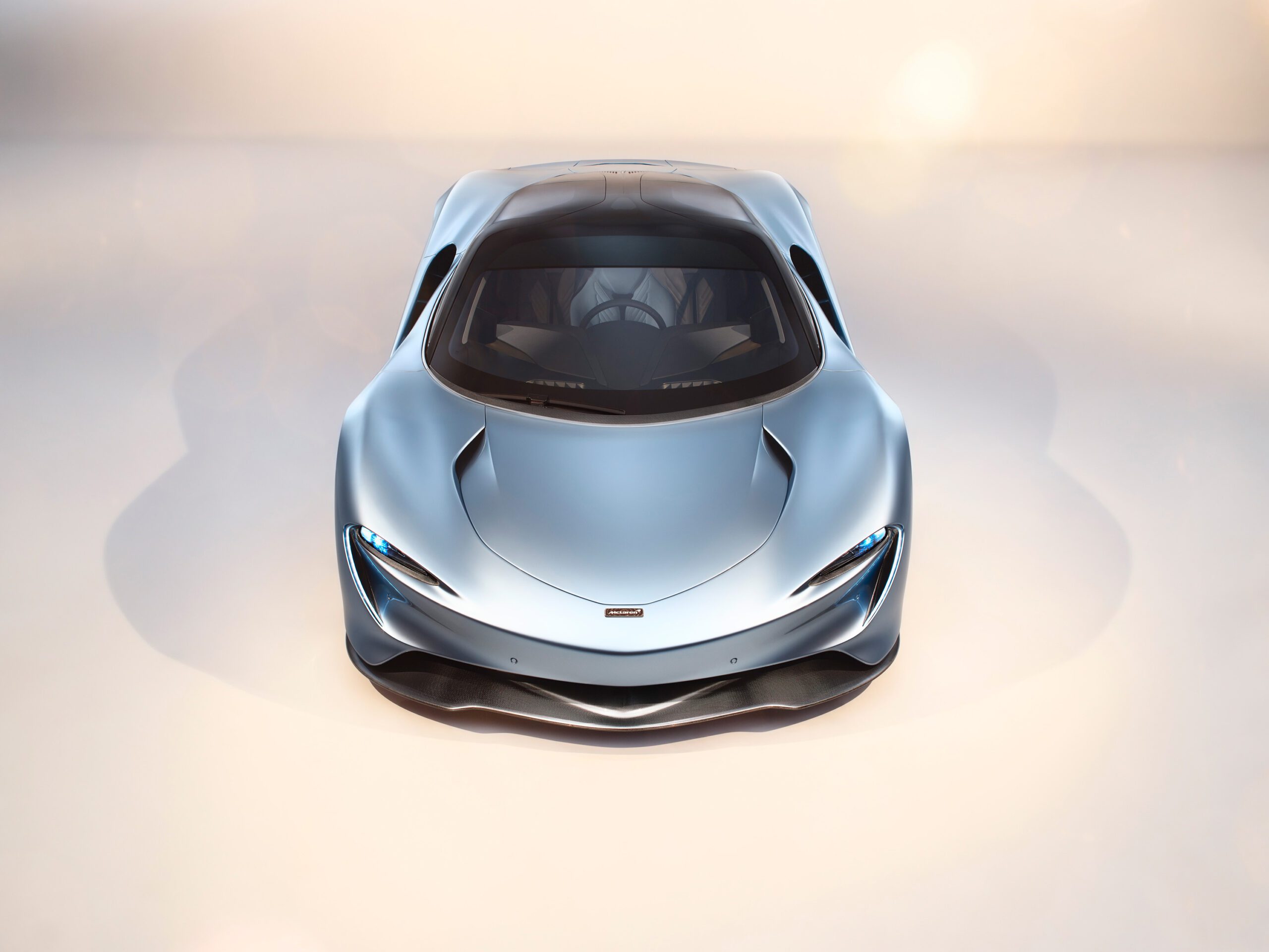 A sleek, silver-blue McLaren Speedtail sports car is pictured from a high front angle. The car features a streamlined, aerodynamic design with a prominent front grille, sharp LED headlights, and a smooth, glossy finish expertly highlighted by a talented car retoucher. The background is a soft, blurred gradient of light tones.