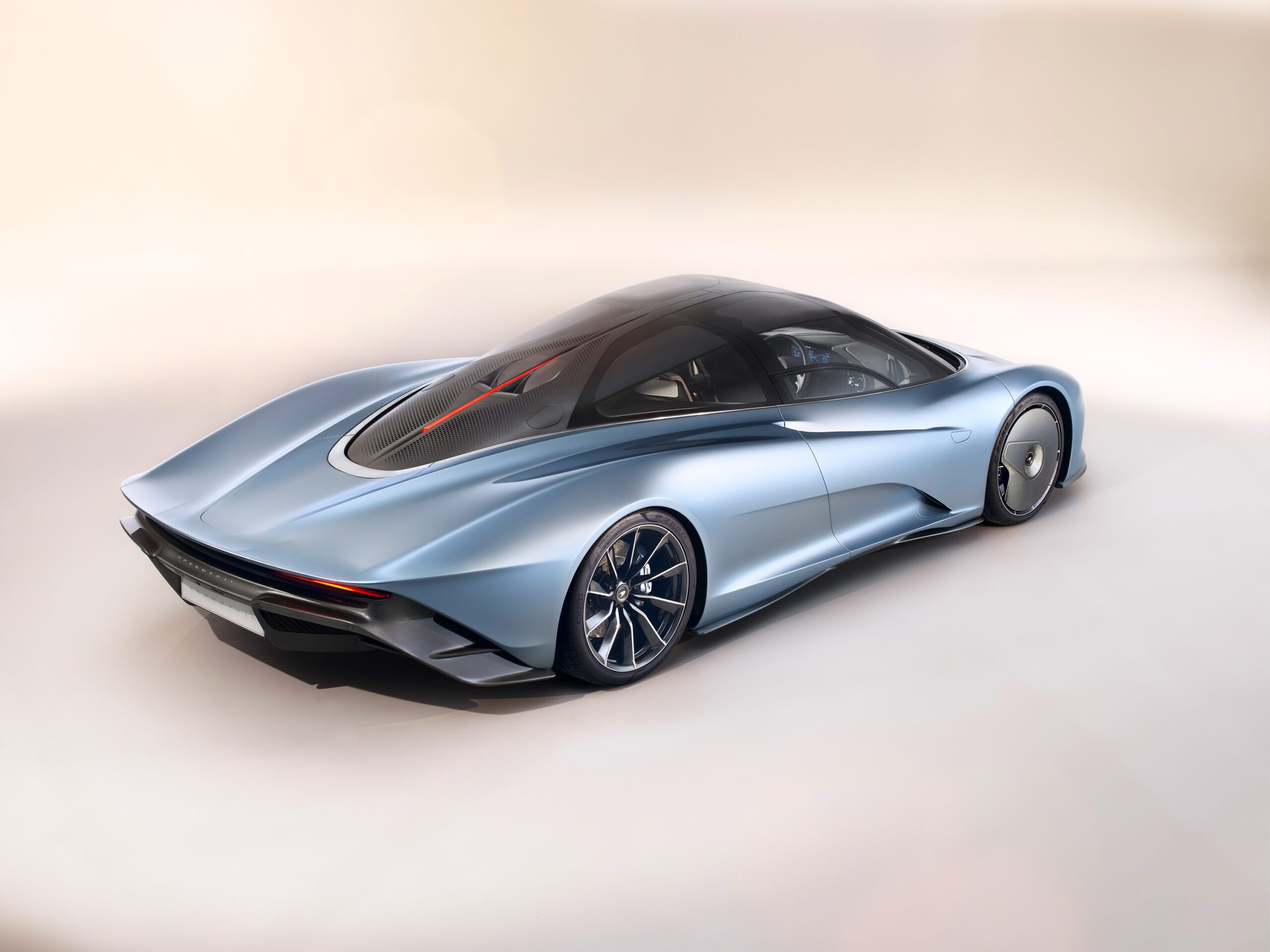 A sleek, futuristic McLaren Speedtail in silver-blue boasts a smooth aerodynamic design, black-tinted roof, streamlined body, and intricate wheel designs. Expertly presented by a skilled car retoucher against a light gradient background, it showcases a modern high-performance aesthetic.