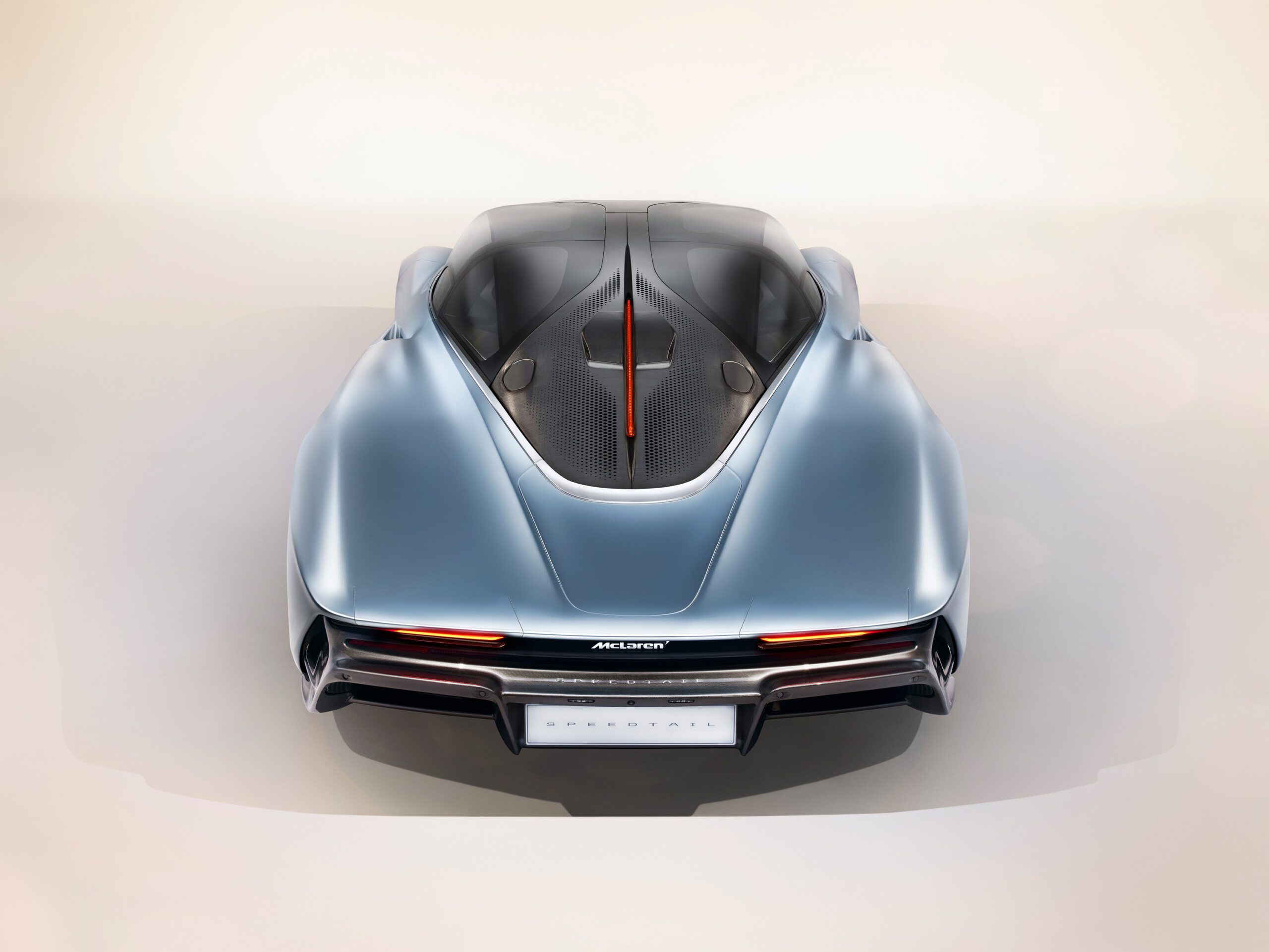 A sleek, silver-blue McLaren Speedtail is viewed from the top, displaying its aerodynamic design. The car features a transparent roof, revealing a portion of the interior and central engine. The streamlined rear end has distinctive taillights, and the license plate reads "Speedtail," captured expertly by a car retoucher.