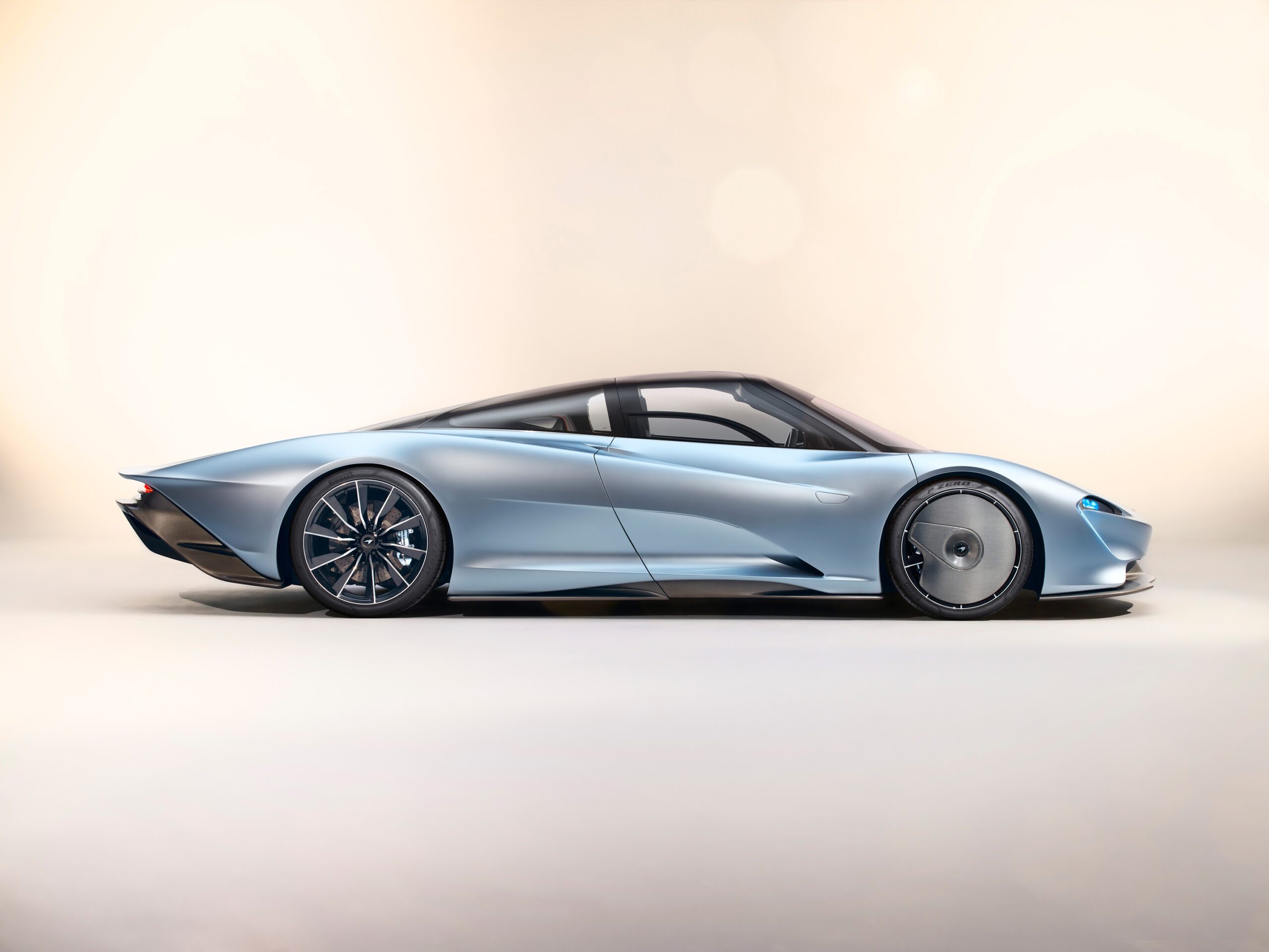 Side view of a sleek, silver-blue McLaren Speedtail with a futuristic design, featuring a low profile, aerodynamic curves, large rear wheels, and slim headlights against a neutral background. Perfectly captured by a skilled car retoucher.