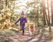 A young girl wearing a purple jacket, knit hat, and red boots is joyfully running on a forest path while holding a leash attached to a happy golden retriever. Sunlight filters through the trees, creating a warm and inviting atmosphere—like an image so perfect it could've been touched by a high-end retoucher.