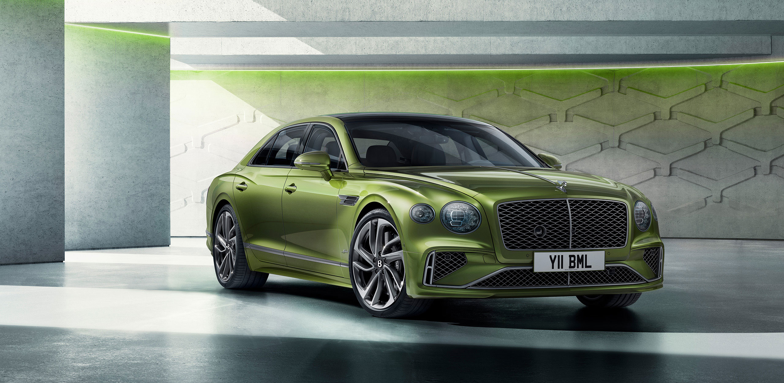 A sleek, green Bentley Flying Spur Speed with a prominent grille and sporty design is parked in a modern, minimalist garage. The car has shiny alloy wheels and the license plate reads "Y11 BML." The background features a geometric wall with subtle lighting accents, resembling a CGI render.