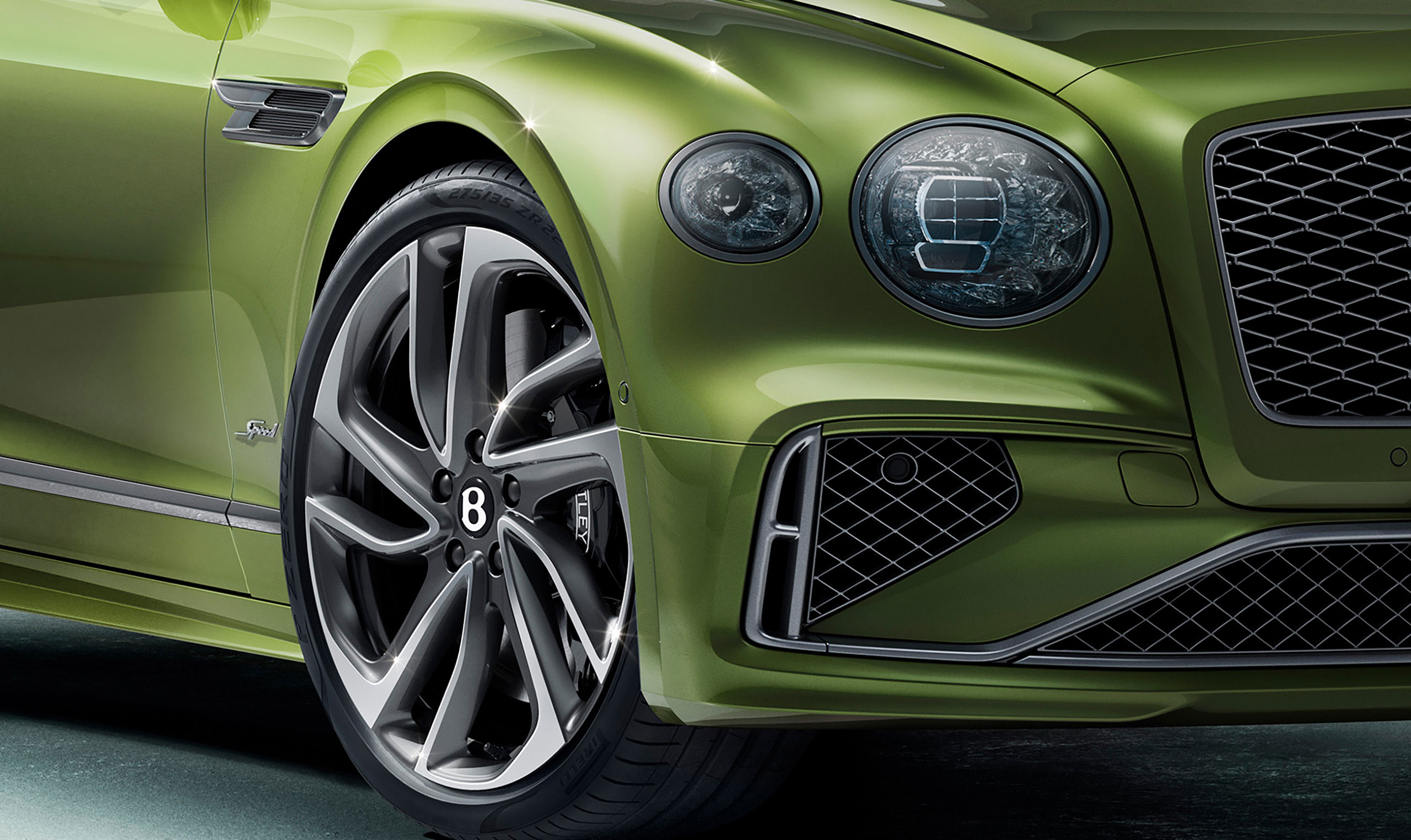 Close-up of the front section of a green Bentley Flying Spur Speed, showcasing a sleek, angular headlight, a prominent front grille with chrome trim, and a multi-spoke alloy wheel featuring the manufacturer's emblem. The car's metallic paint reflects the light subtly in this CGI rendering.