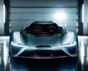 A sleek, futuristic blue Nio EP9 is showcased in a well-lit garage. The car features sharp, angular headlights and a low, aerodynamic design. Bright lights in the background illuminate the scene, reflecting off the high end retoucher's meticulous work on its smooth surface.