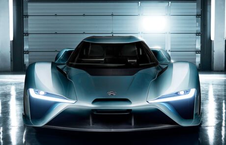 A sleek, futuristic blue Nio EP9 is showcased in a well-lit garage. The car features sharp, angular headlights and a low, aerodynamic design. Bright lights in the background illuminate the scene, reflecting off the high end retoucher's meticulous work on its smooth surface.