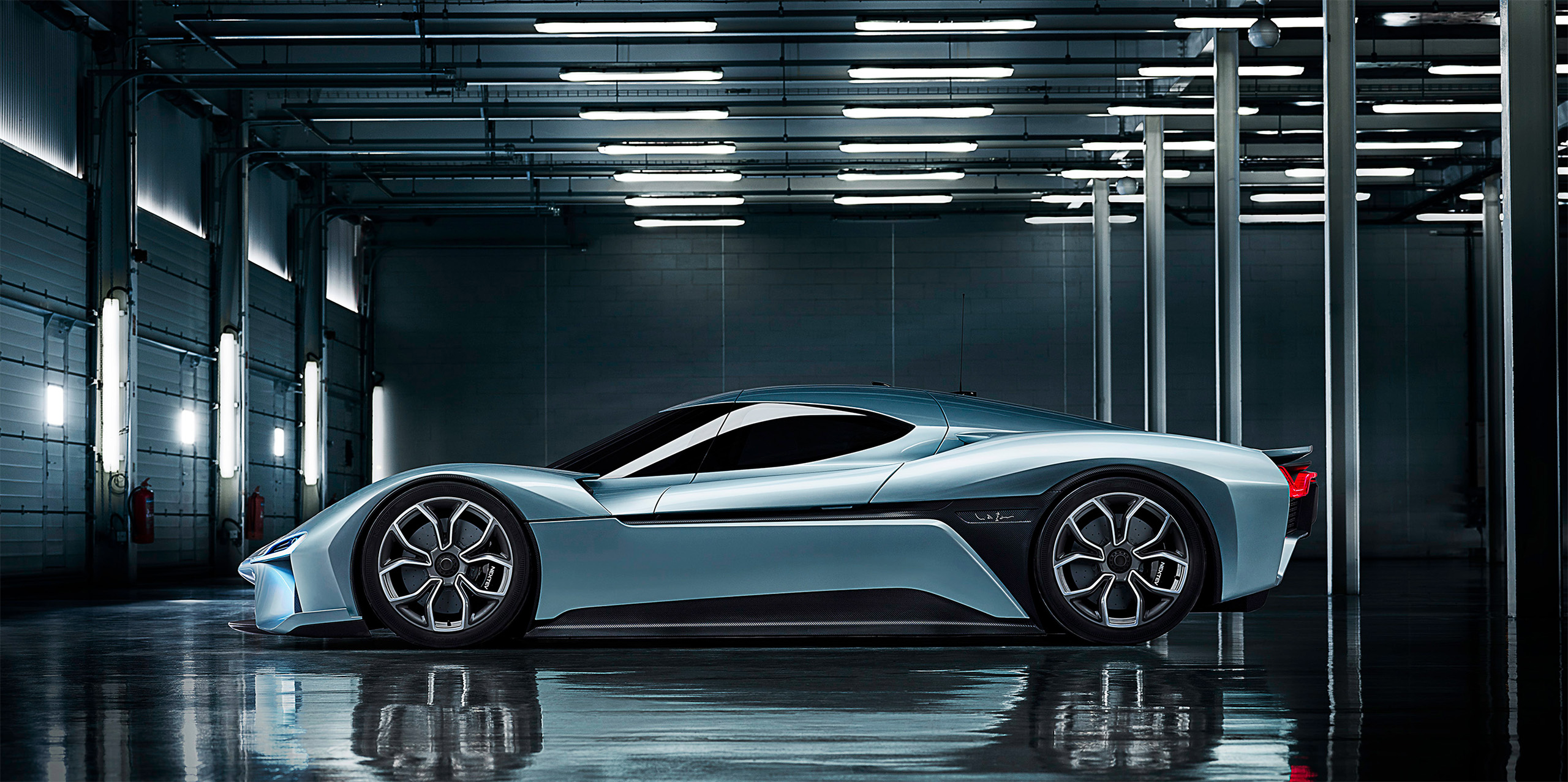 A sleek, futuristic silver-blue sports car is parked in a modern industrial garage. The car features a low profile, aerodynamic design, and large wheels. The garage has bright overhead lights and metallic pillars, reflecting on the polished floor.