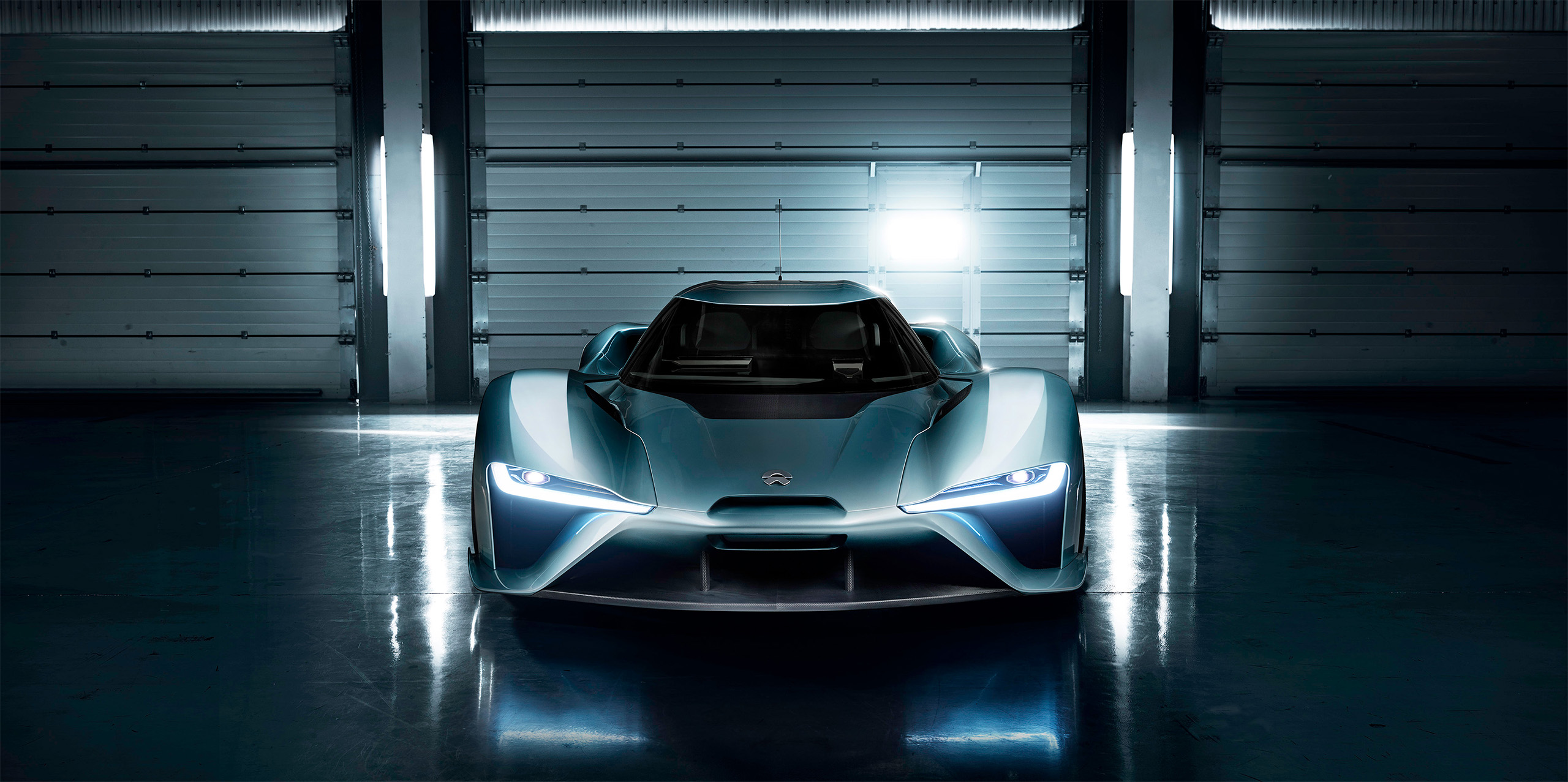 A sleek, futuristic blue sports car with glowing headlights is showcased in a dimly lit, modern industrial garage. The car is positioned front and center, highlighting its aerodynamic design and aggressive styling.