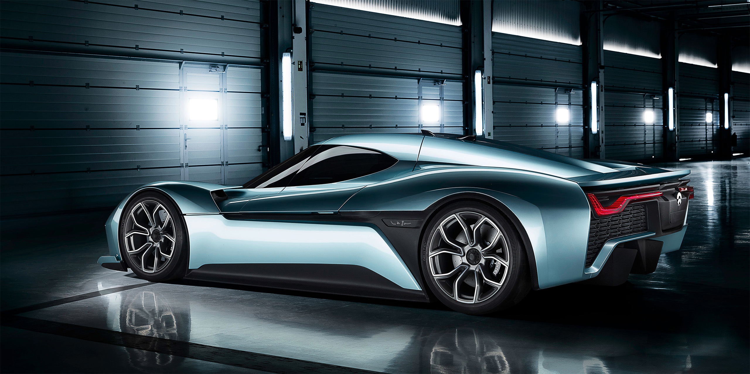 A sleek, futuristic electric sports car with a metallic blue finish is parked in a dimly lit, industrial-style garage with large doors and bright overhead lights. The car's aerodynamic design and large wheel rims are prominently visible.