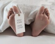 Close-up of two bare feet protruding from white bedcovers, with a tag attached to one toe reading "STAY HOME. SAVE LIVES." The scene evokes a stark message related to the covid19 pandemic—staying home protects health and safety.