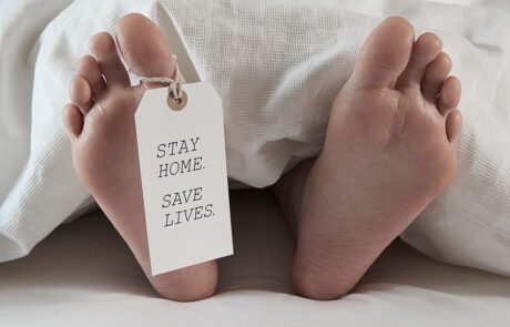 Close-up of two bare feet protruding from white bedcovers, with a tag attached to one toe reading "STAY HOME. SAVE LIVES." The scene evokes a stark message related to the covid19 pandemic—staying home protects health and safety.