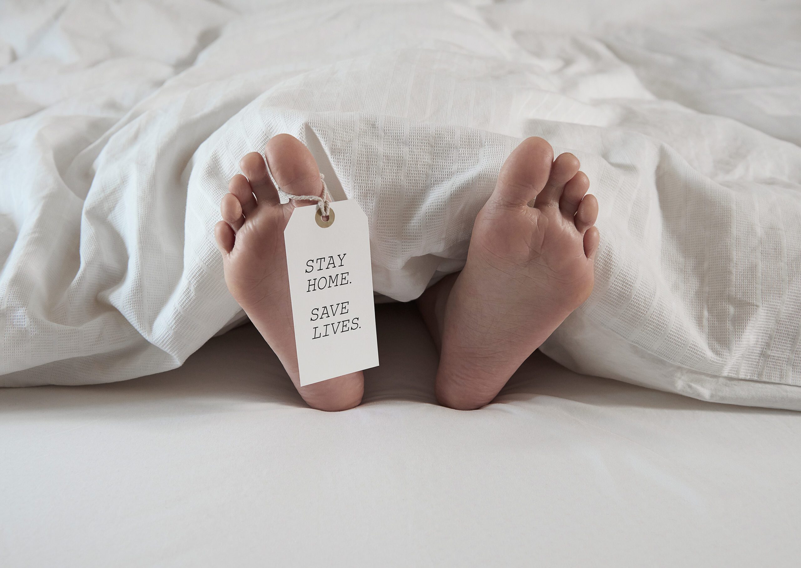 Two bare feet protrude from under a white blanket with a tag attached to the left foot. The tag reads "STAY HOME. SAVE LIVES." The scene starkly highlights the impact of COVID-19 and underscores the importance of staying home to protect public health during the pandemic.