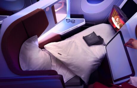 A luxurious Virgin Atlantic A350 Airbus business class seat converted into a flatbed with white bedding. A pair of glasses and a pouch rest on the side table. The screen displays a cartoon, and a person's hand is visible adjusting the tray table. Soft ambient lighting sets a cozy mood.