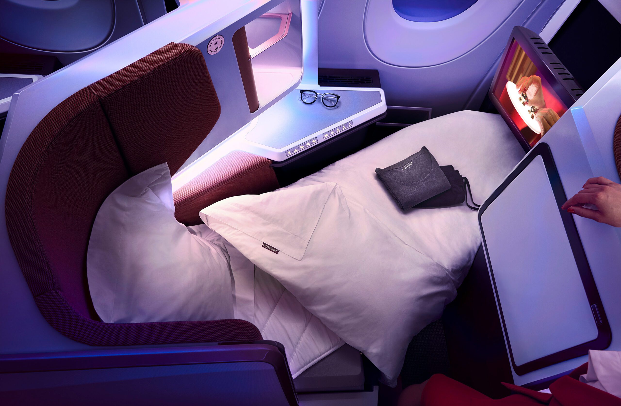 A luxurious Virgin Atlantic A350 Airbus business class seat converted into a flatbed with white bedding. A pair of glasses and a pouch rest on the side table. The screen displays a cartoon, and a person's hand is visible adjusting the tray table. Soft ambient lighting sets a cozy mood.