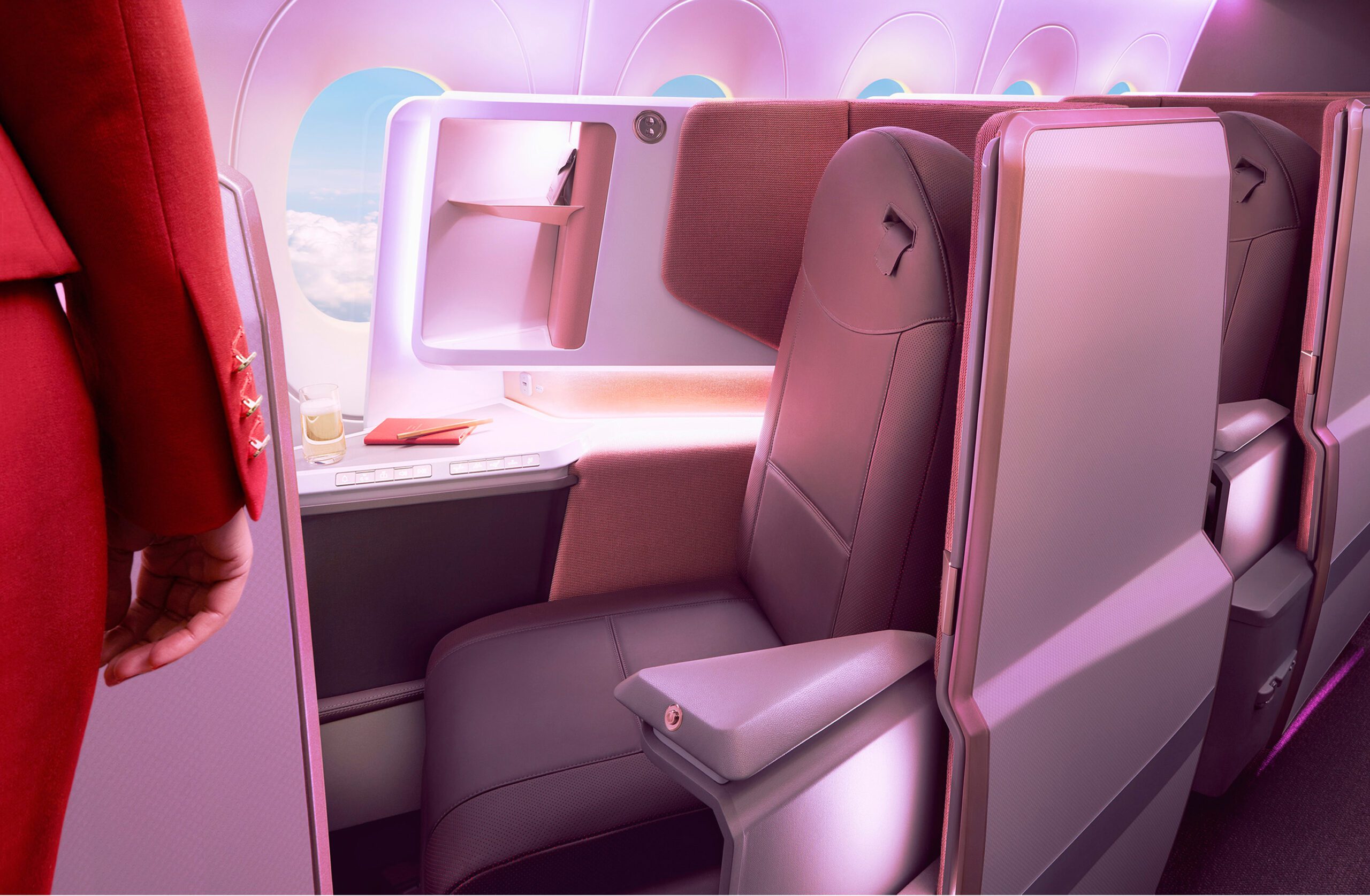 The image shows a luxurious first-class airplane seat on Virgin Atlantic's A350 Airbus. The seat is spacious with ample legroom, upholstered in dark leather, and has a privacy partition. A flight attendant in a red uniform stands beside the seat, which is captured under soft, pink lighting through professional retouching.