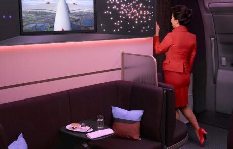 A flight attendant in a red uniform stands near a curtain in the modern Virgin Atlantic A350 Airbus cabin, looking at a screen showing an aerial view. The cabin features plush seating, pillows, a small table with snacks and drinks, and ambient lighting with star-like designs, all resembling professional retouching.