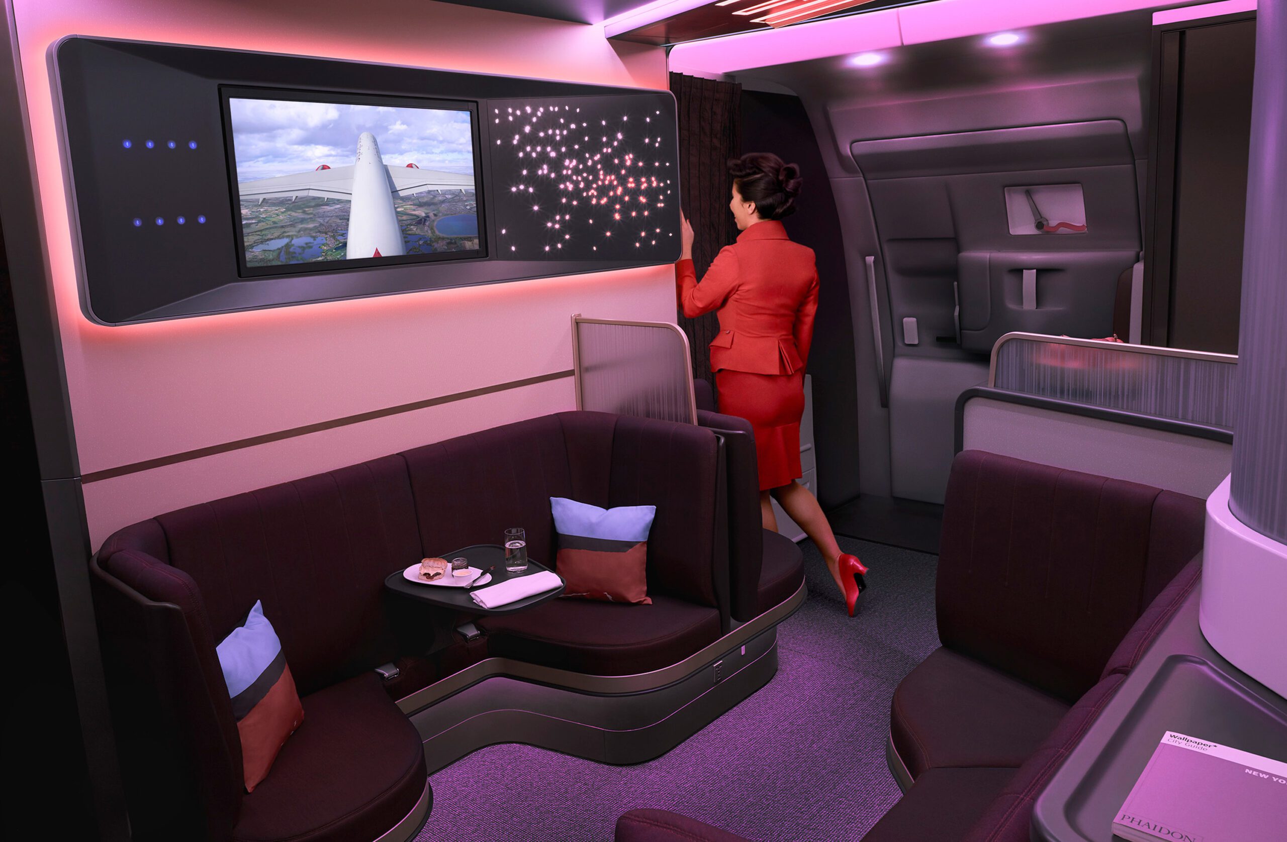 A flight attendant in a red uniform stands facing a door in the Virgin Atlantic A350 Airbus cabin. The space features a lounge area with a screen showing an aerial view, L-shaped seating with pillows, and a small tray with food and drink. The modern lighting casts a purple hue, enhancing the scene with professional retouching.