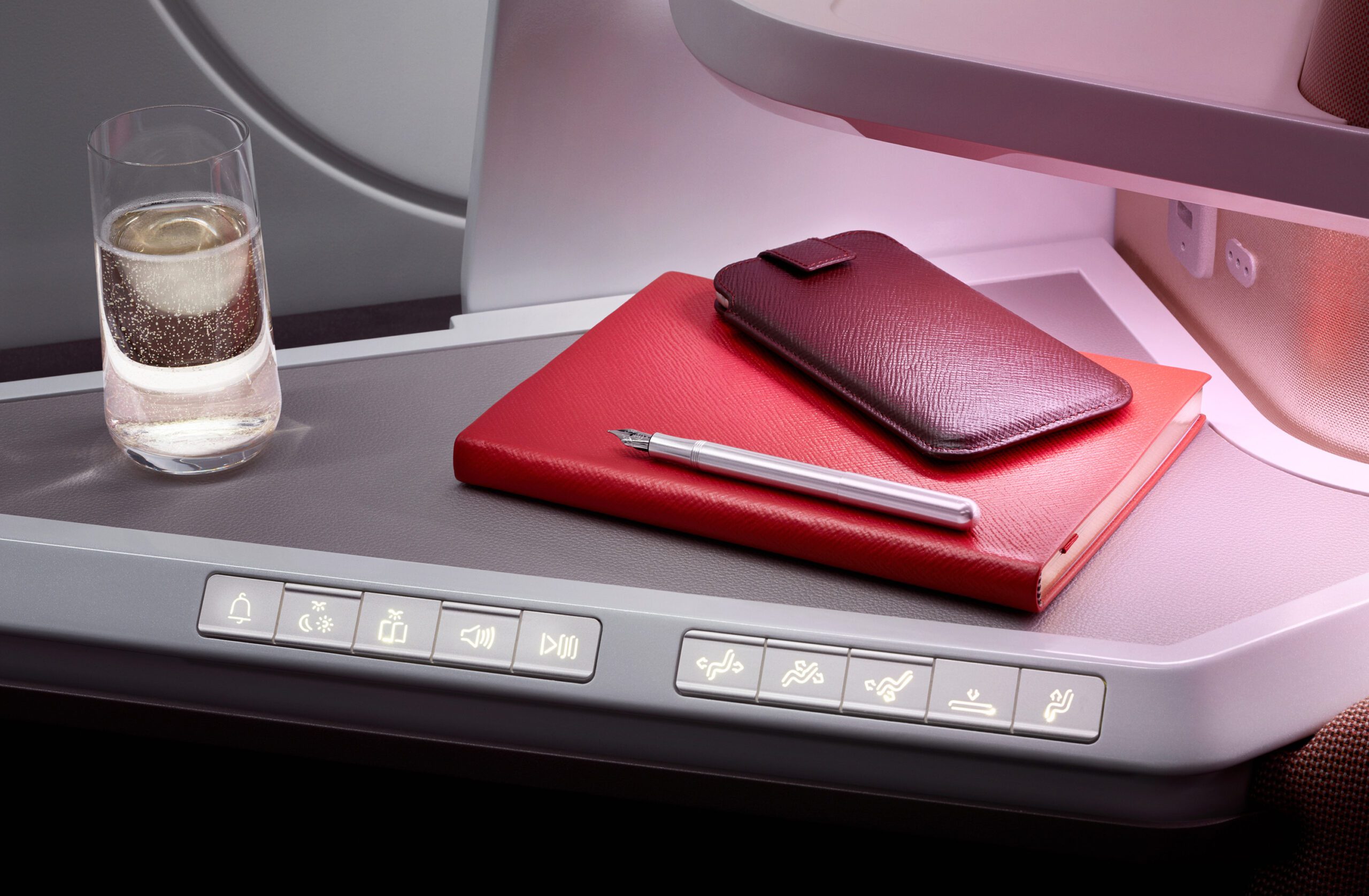 A close-up of an airplane tray table in a Virgin Atlantic A350 Airbus premium cabin. On the table are a glass of water, a red leather notebook, a matching red leather case, and a silver pen. The tray features various function buttons along the bottom edge for light and seat adjustment.