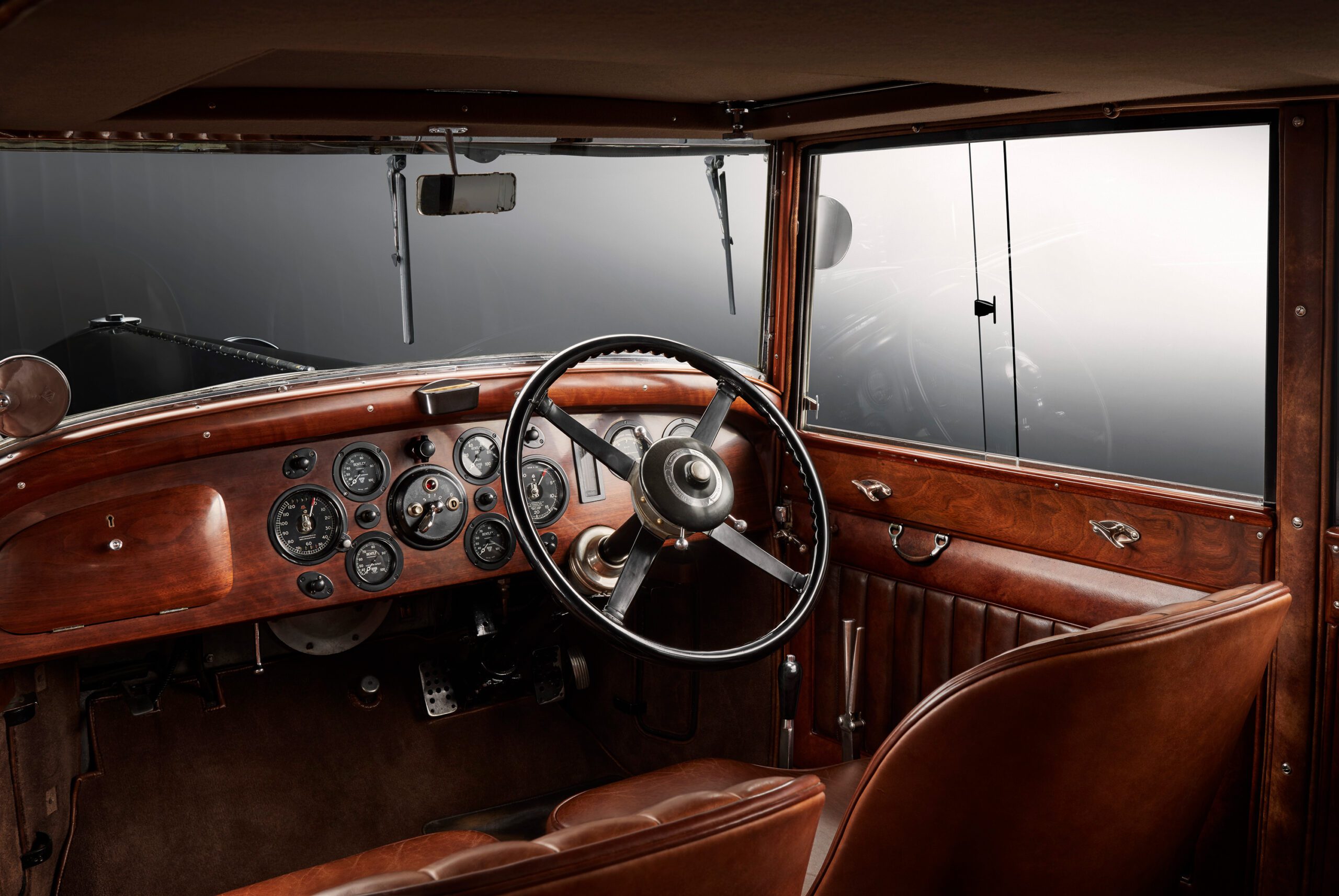 WO Bentley's own car interior featuring a large steering wheel, classic gauges, and wooden dashboard. The seats and door panels are upholstered in brown leather. The ambiance is nostalgic with a dark, reflective exterior visible through the window.