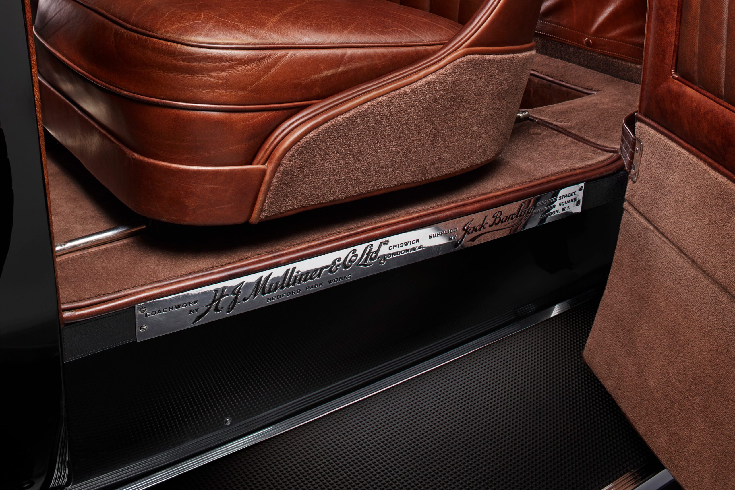 Close-up view of a WO Bentley's interior showing the brown leather seats and carpeted floor. A metallic nameplate with engraved text, including "H.J. Mulliner & Co.," is affixed to the door sill, emphasizing the vehicle's custom craftsmanship.