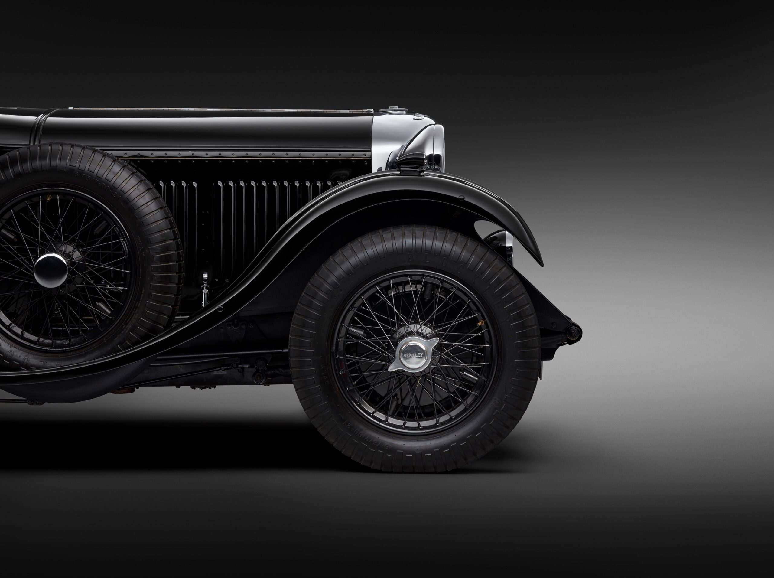 Side view of WO Bentley's own 8 litrecar, showcasing its distinctive rounded fender, exposed chrome-rimmed headlight, black wire-spoked wheel, and detailed tire. The background is a gradient from dark to light grey, emphasizing the car's sleek design.