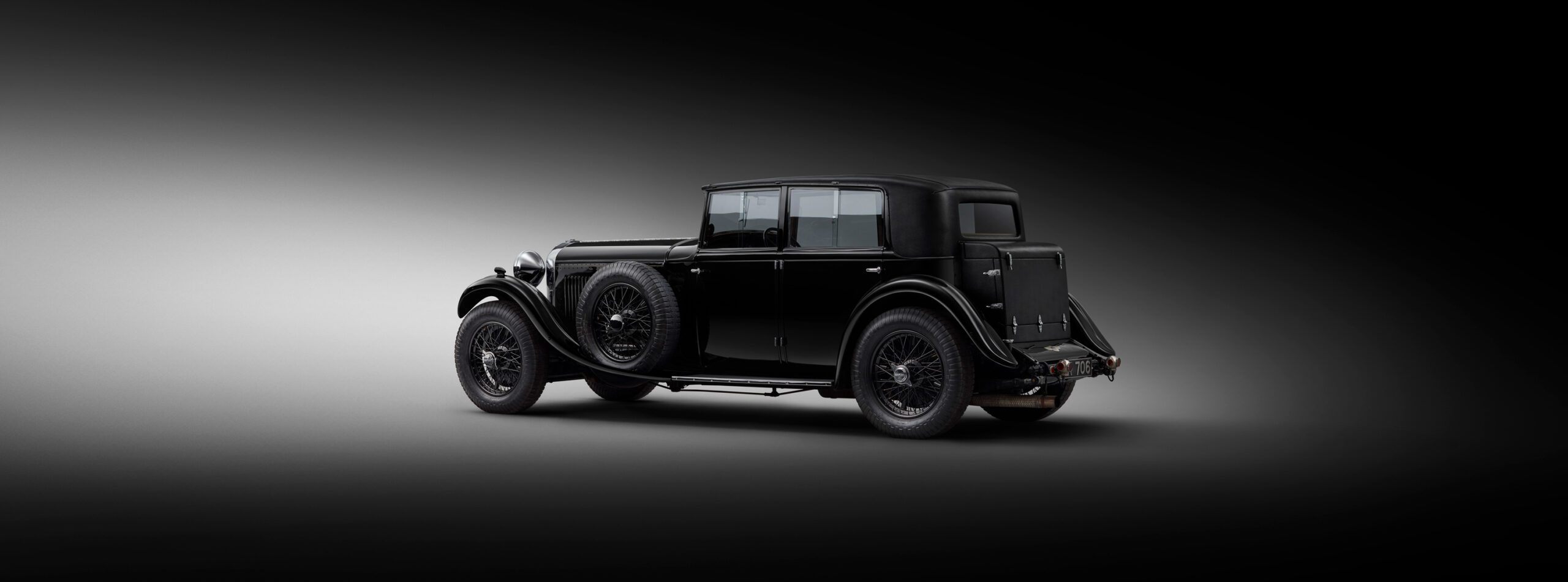 WO Bentley's own 8 litre car with a sleek, glossy finish is parked on a gradient background that transitions from black to gray. The car features large spoked wheels, two spare tires mounted on the sides, and an elegant, boxy body design from an early 20th-century era.