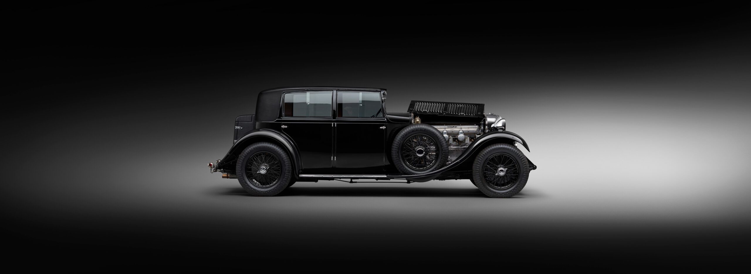 A side view of WO Bentley's own 8 litre car against a gradient black-to-gray background. The car features a sleek, elongated body, round headlights, and an exposed engine on the front right side, giving it a classic and elegant appearance.