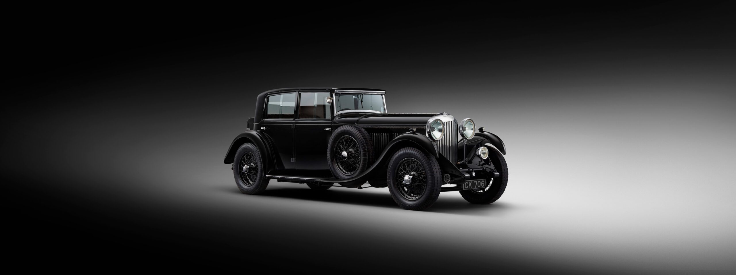 WO Bentley's own 8 litre car is displayed against a dark gradient background. The vehicle has a classic design with round headlights, a long hood, and spoked wheels. The car exudes elegance and showcases a bygone era of automotive craftsmanship.