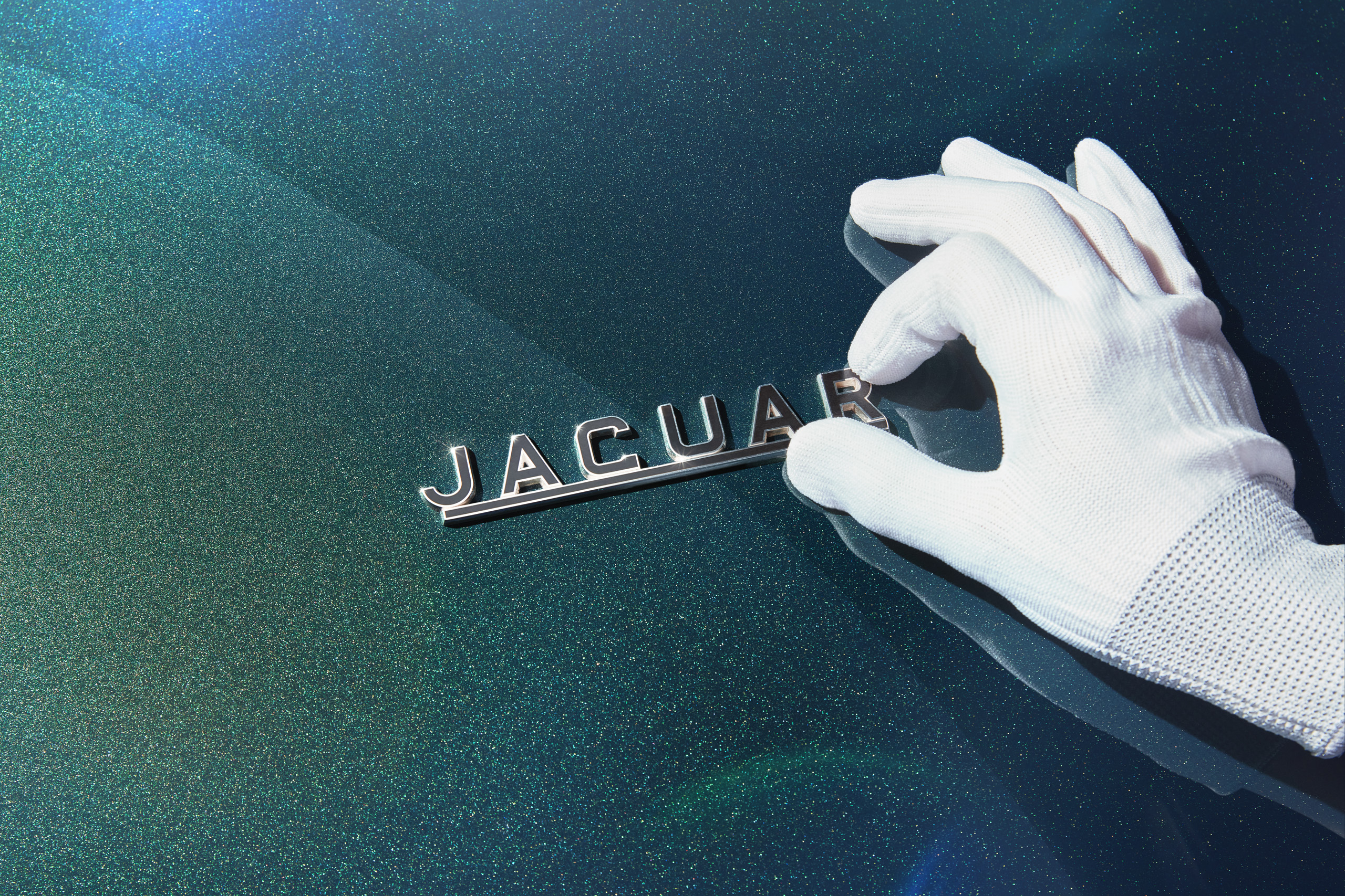 A gloved hand polishes the chrome Jaguar logo on the dark green, metallic paint of a car. The background shows a glimmering finish, highlighting the sleek, luxurious emblem.