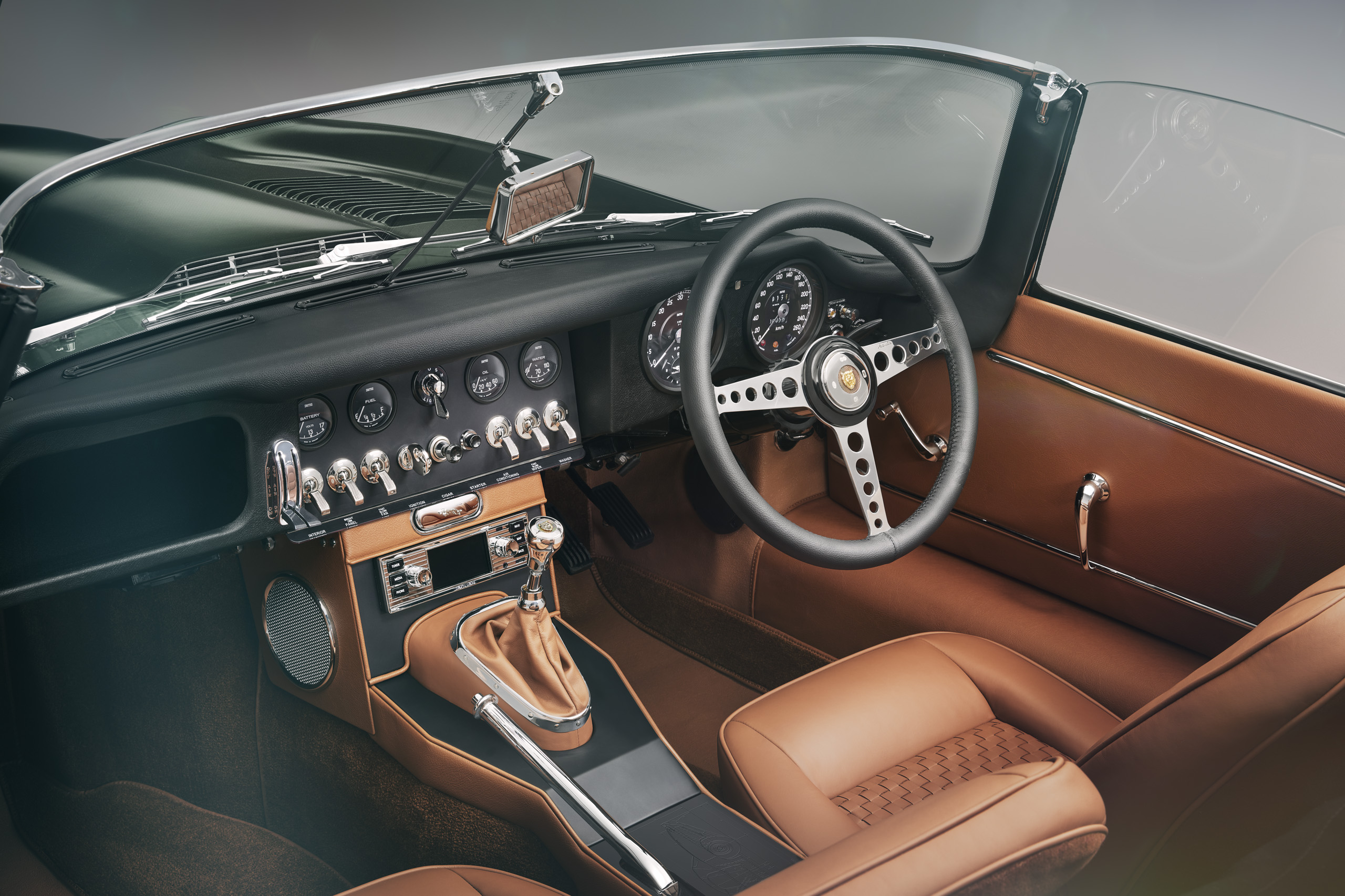 Interior of a vintage sports car with a brown leather dashboard and seats. The steering wheel is black with a metallic center, surrounded by an array of analog gauges and switches. The gear shift is covered with a brown leather boot.