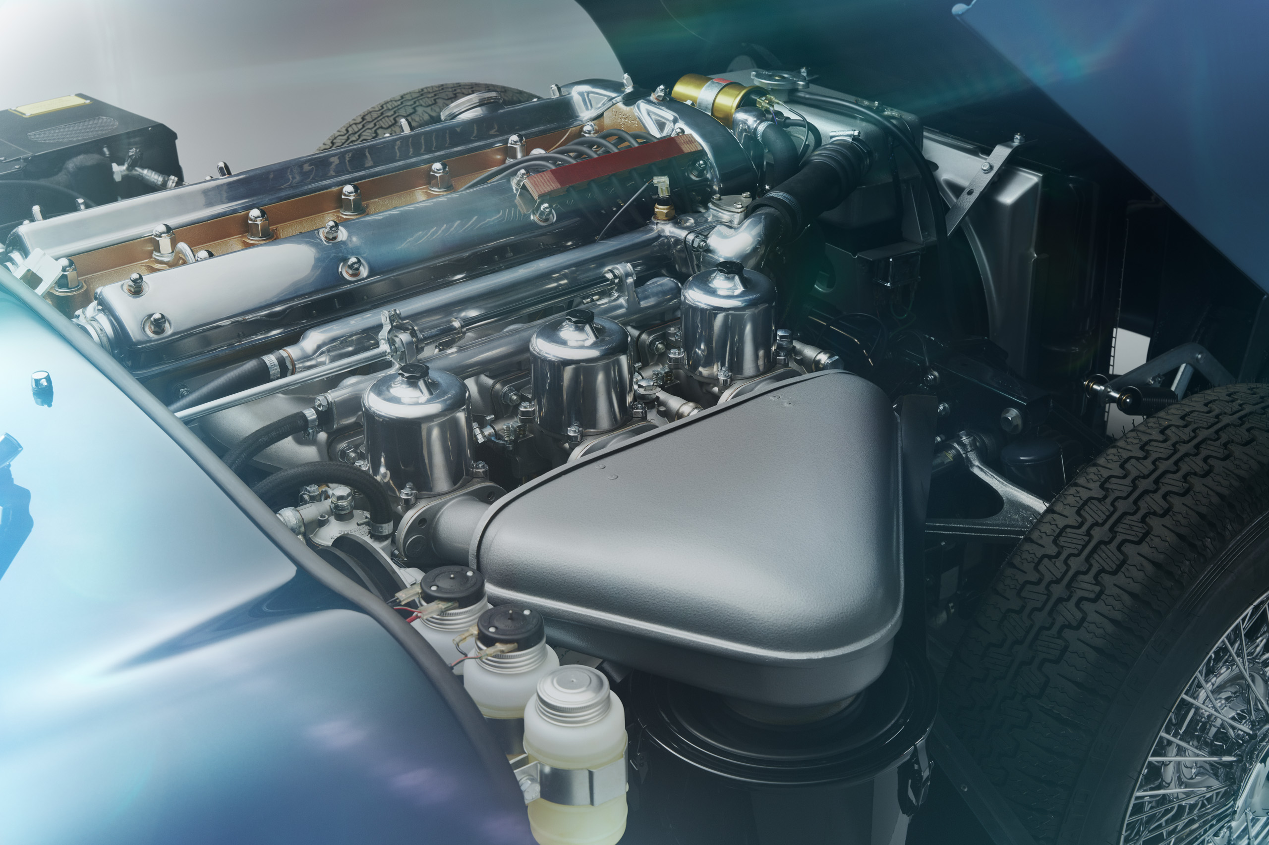 Close-up of a vintage car engine with a polished metal finish. The image shows intricate components including carburetors and a sleek air filter, surrounded by various hoses and parts. Sunlight creates lens flare on the metallic surfaces.