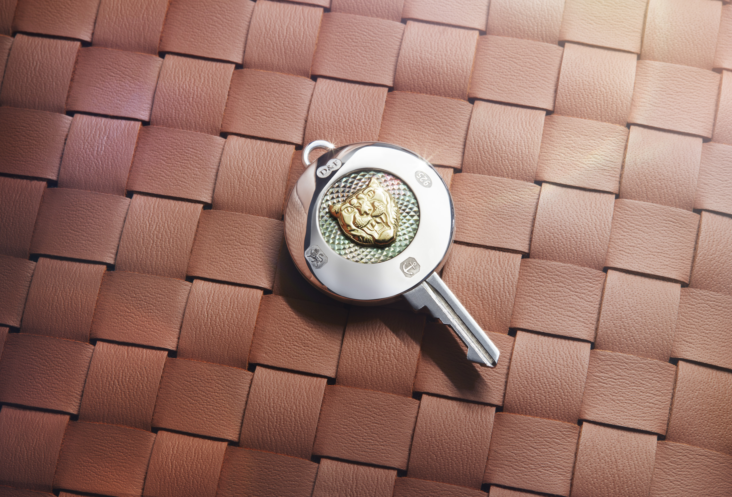 A silver key with a decorative gold emblem on its handle, featuring an intricate design, rests on a woven leather surface in shades of brown.