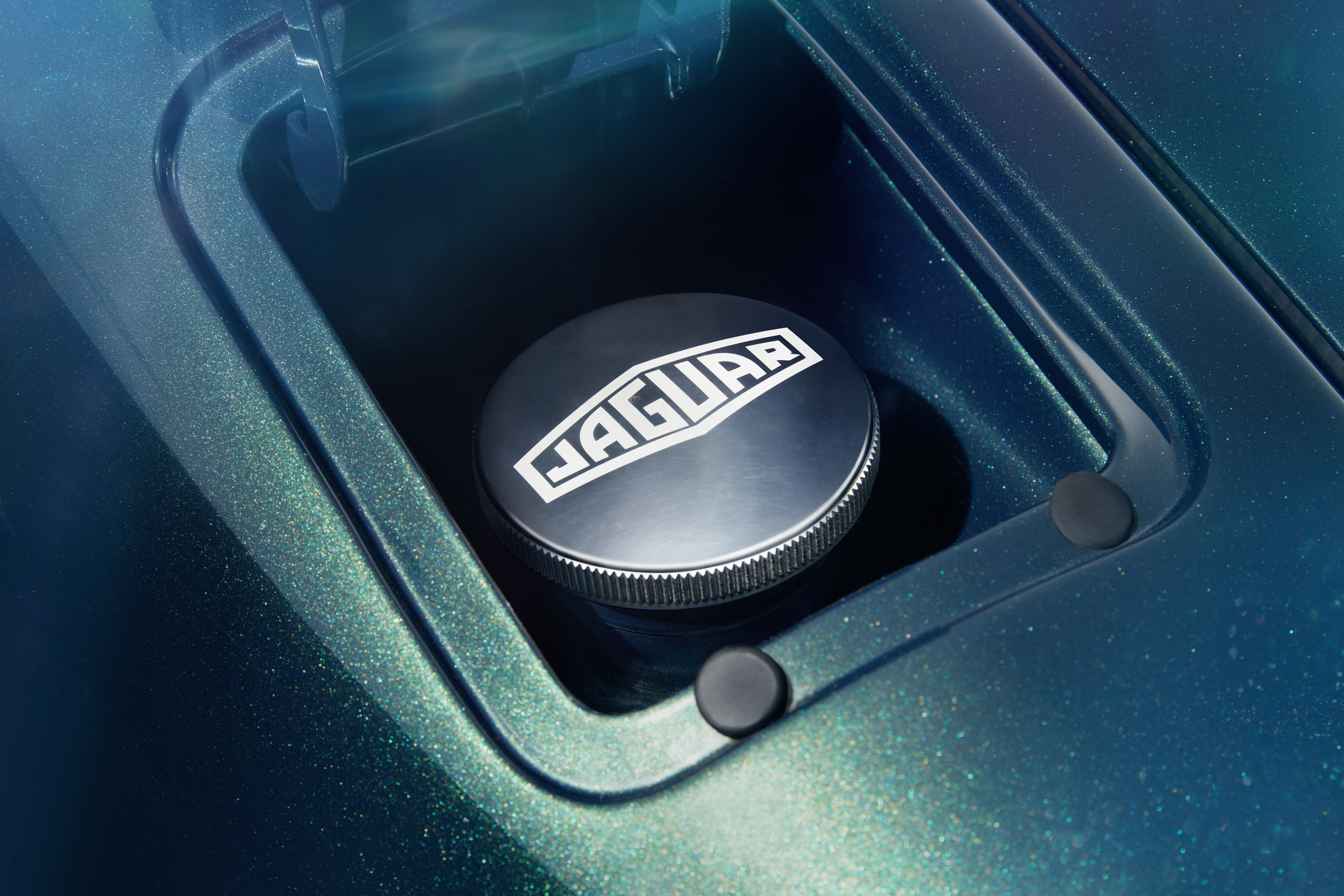 Close-up of a metallic fuel cap with a Jaguar logo, set in a recessed compartment on a sleek, metallic blue-green vehicle surface. The area is well-lit, highlighting the texture and details.