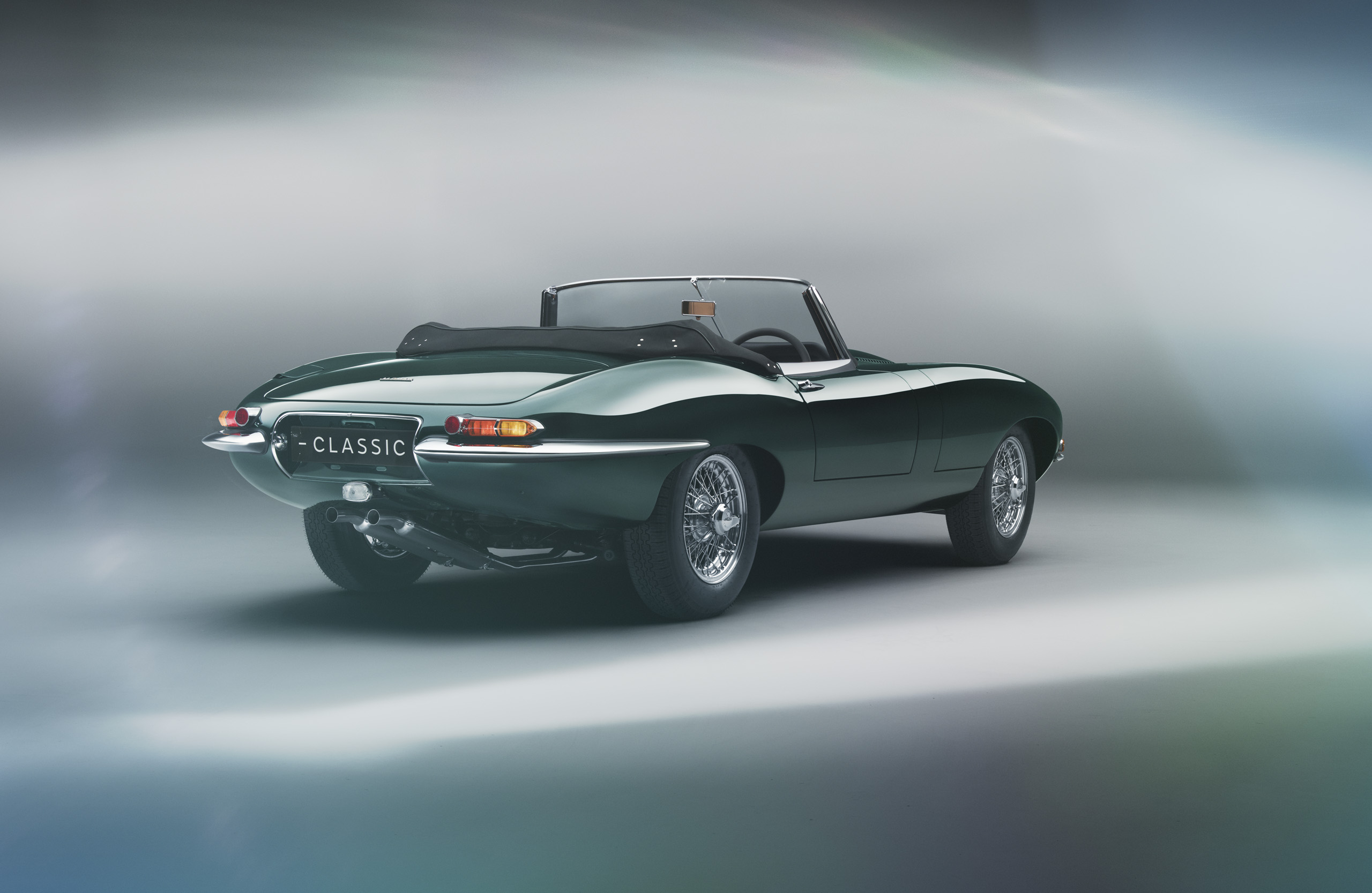 A classic green convertible sports car with sleek curves is showcased against a blurred, light-infused background. The license plate reads "CLASSIC." The car's top is down, highlighting its elegant design and vintage appeal.