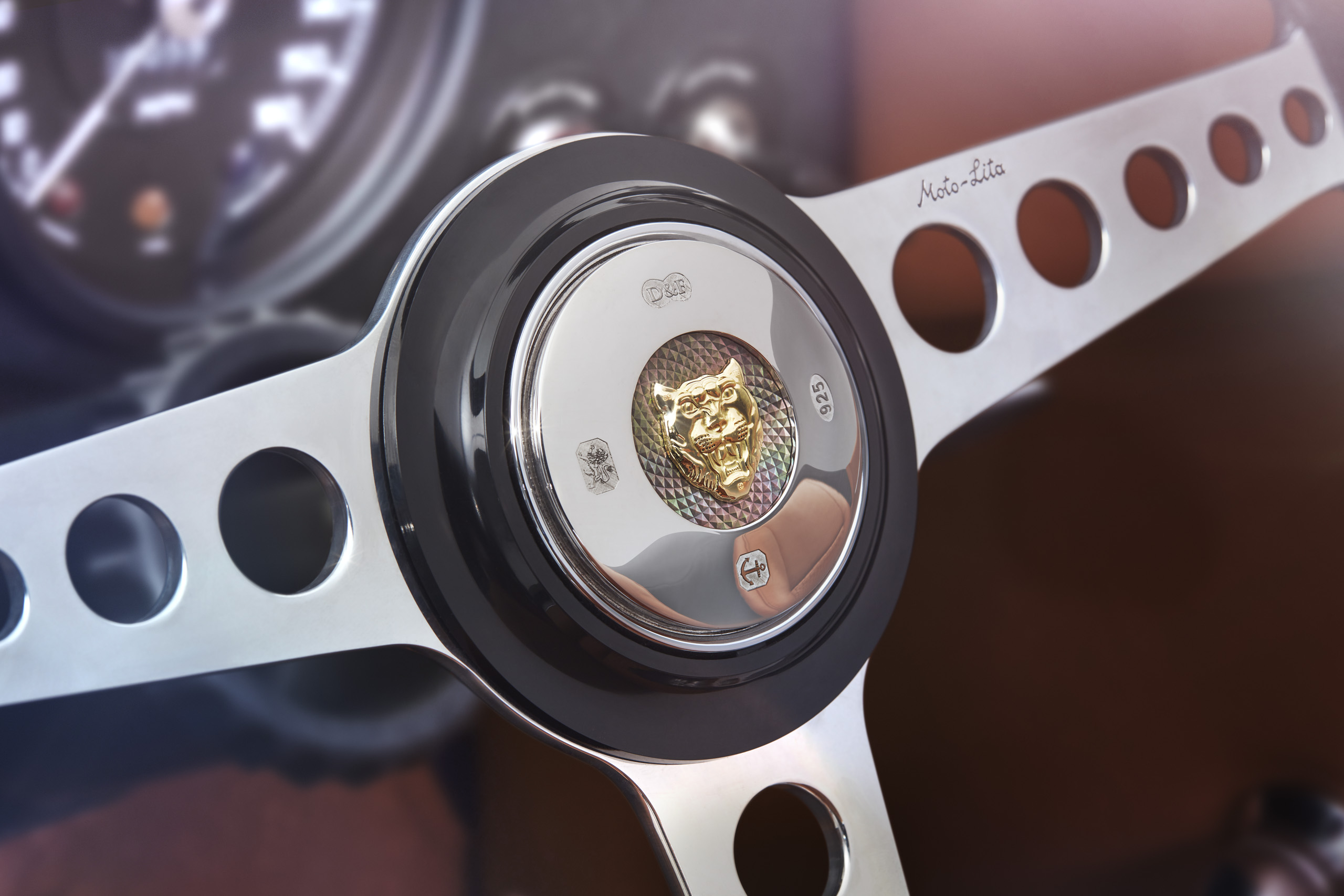 Close-up view of a classic car steering wheel with a metallic design. The center features an emblem of a golden jaguar face. The wheel has circular cutouts on the spokes and an instrument panel is slightly visible in the blurred background.