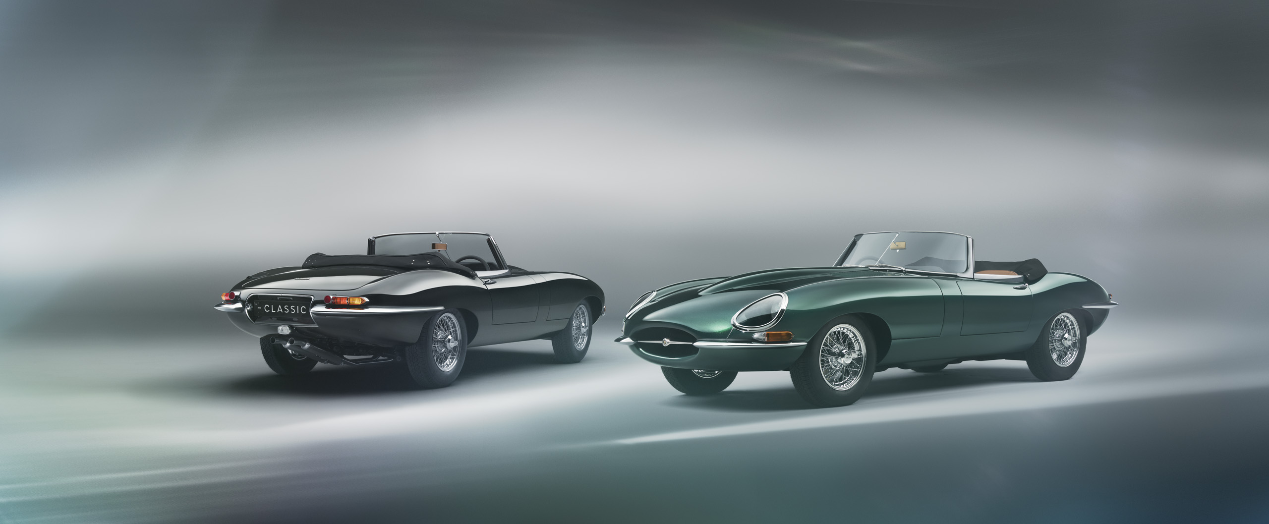 Two classic convertible cars with sleek, curved designs. One is black, seen from behind, and the other is dark green, viewed from the front. Both cars are set against a blurred, gradient background.