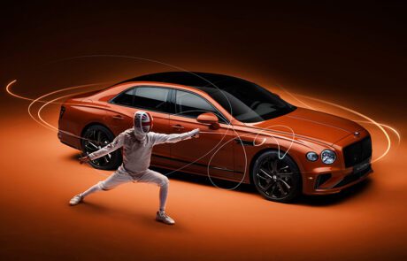 A fencer in full gear strikes a pose, with a sleek, orange luxury car in the background. The scene is dynamically lit, with motion trails accentuating the action, set against a warm, dark backdrop.