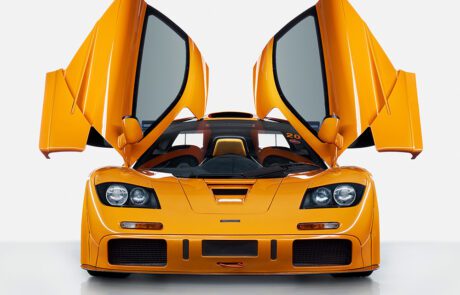 A bright orange sports car with its scissor doors open, resembling wings. The car features a sleek design with large air intakes and distinctive angular headlights, set against a plain white background.