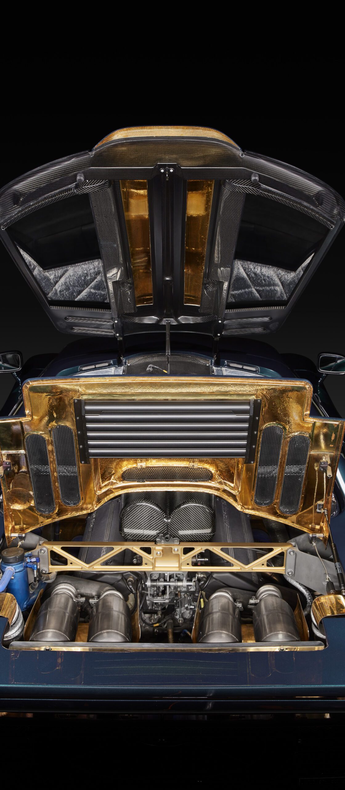 A close-up view of a luxury sports car's rear hood open, revealing a gold-accented, high-performance engine with visible exhaust components and intricate mechanical details.