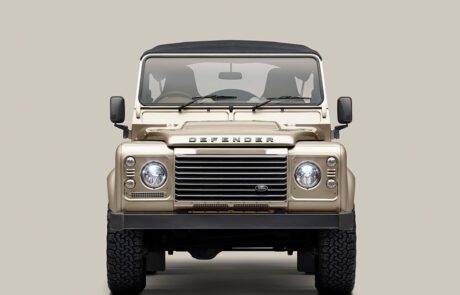 Front view of a beige Land Rover Defender with a black roof, seen against a plain light gray background. The vehicle’s headlights, grille, and rugged tires are clearly visible.
