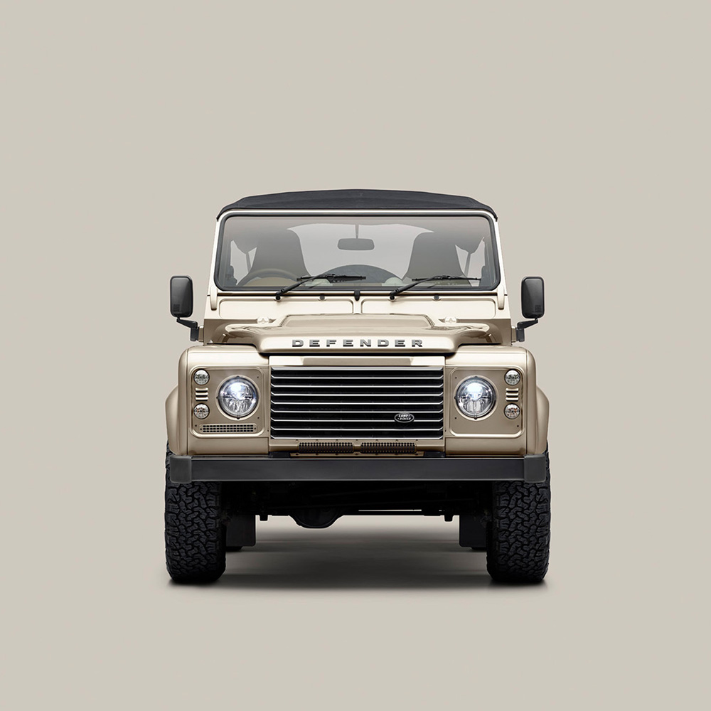 Front view of a beige Land Rover Defender with a black roof, seen against a plain light gray background. The vehicle’s headlights, grille, and rugged tires are clearly visible.
